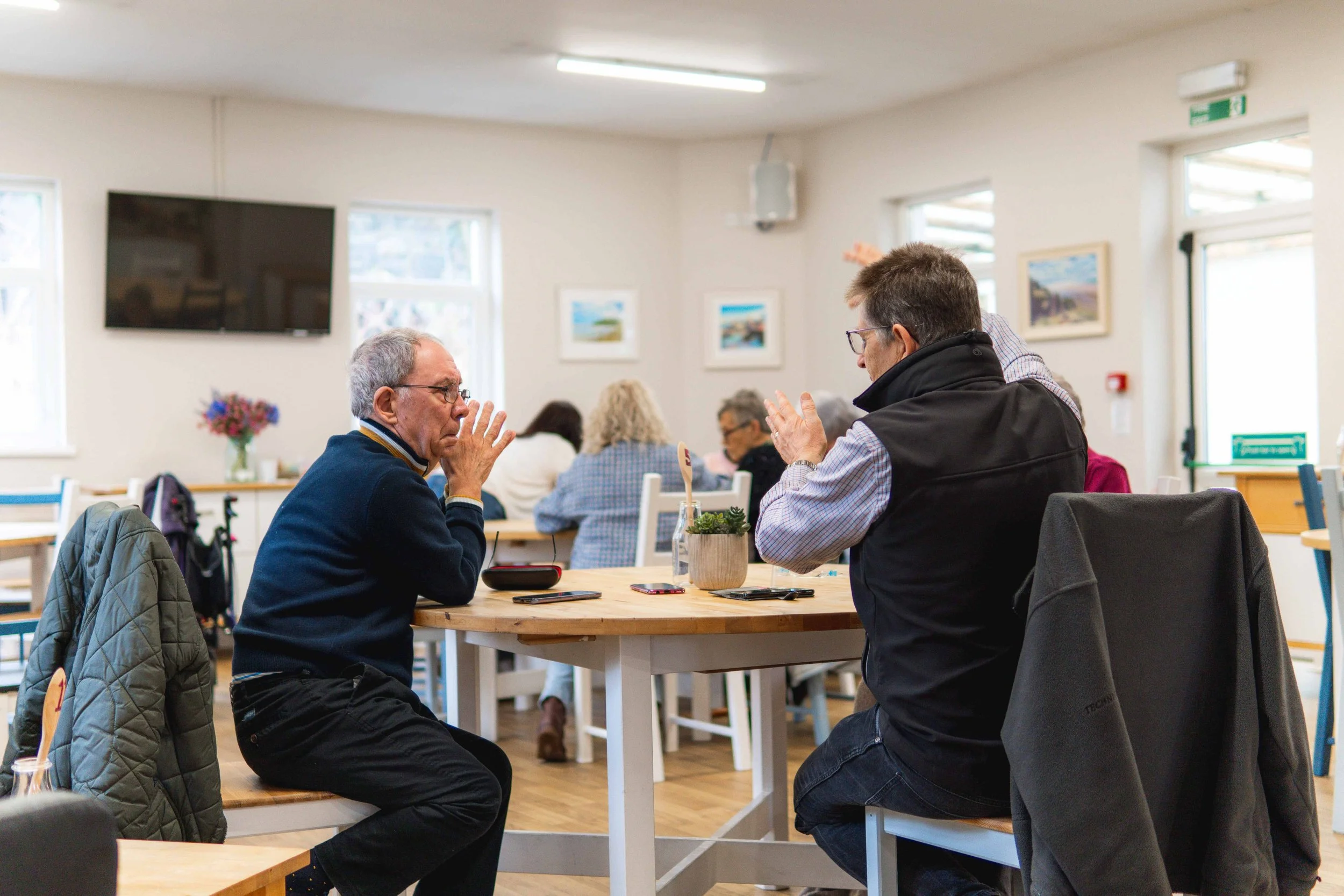 Two men are sitting across from each other at a wooden table in a bright, busy café or restaurant, engaged in a conversation with hand gestures. Other patrons are seated at tables in the background, and there are framed pictures on the walls and a TV screen mounted high.