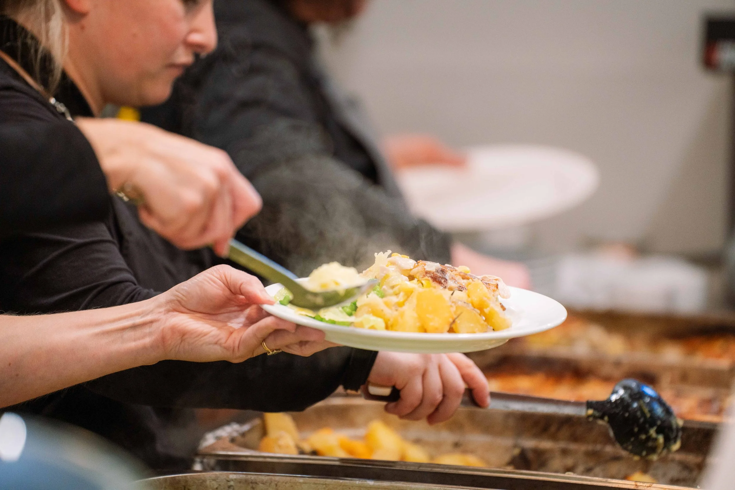 People serving themselves food at a buffet, with one person placing a serving of potato casserole on a plate.