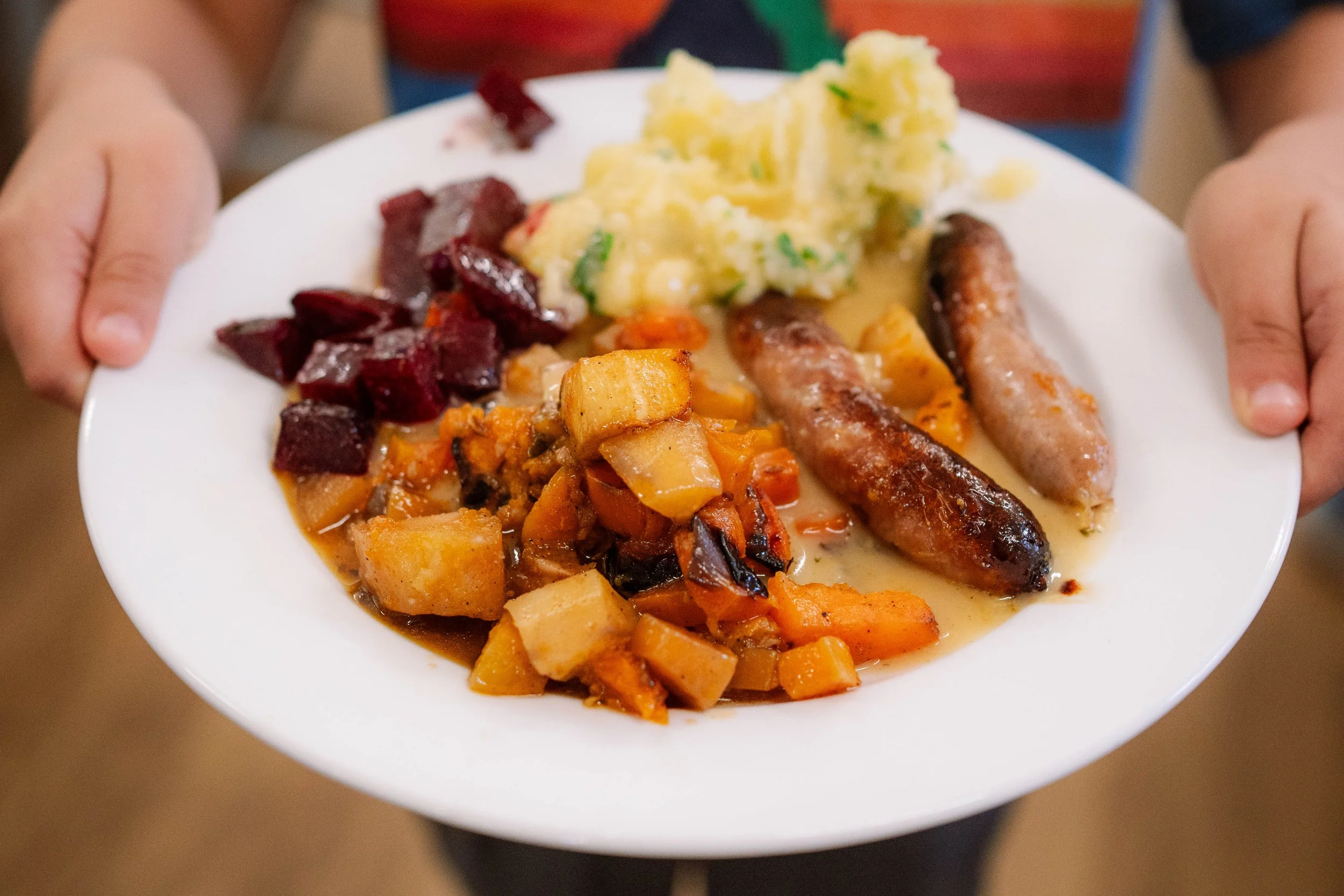 A white plate with mashed potatoes, roasted beets, a vegetable stew, and two sausages being held by a person.