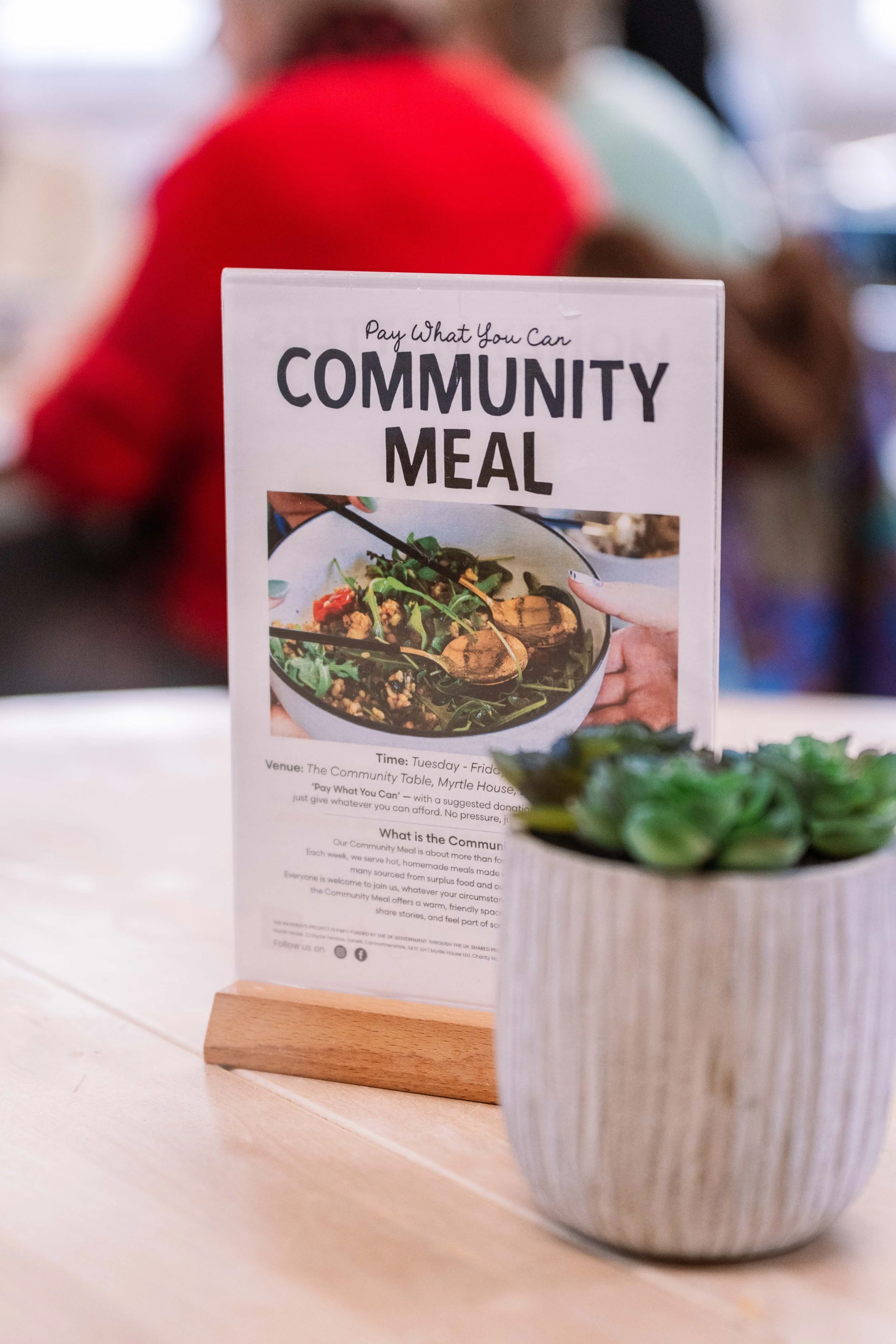 A sign on a table reads 'Pay What You Can Community Meal' with details about the venue and time. The sign features a photo of a bowl of salad or soup. In the foreground, there is a potted succulent plant. In the blurred background, people are present, with one person in a red shirt.