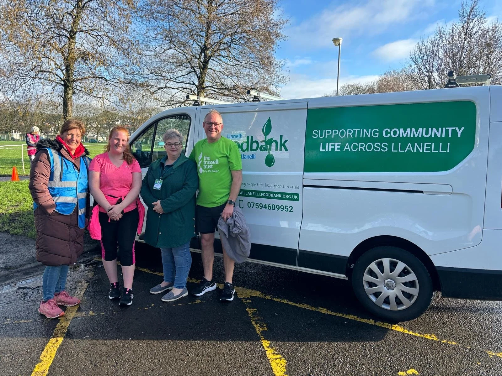 Four people standing outdoors near a parked food bank van, with a park and leafless trees in the background. The van has a green and white sign indicating it supports community life across Llanelli.