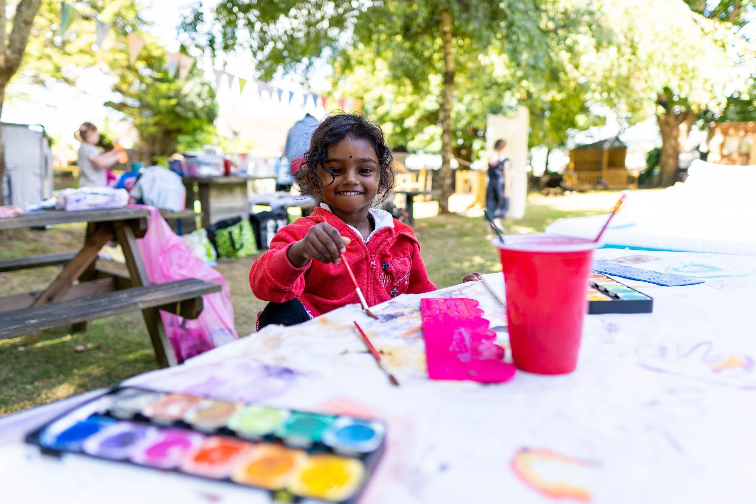 Young girl in red jacket smiling while painting watercolor at outdoor art activity table, with other children and trees in background.