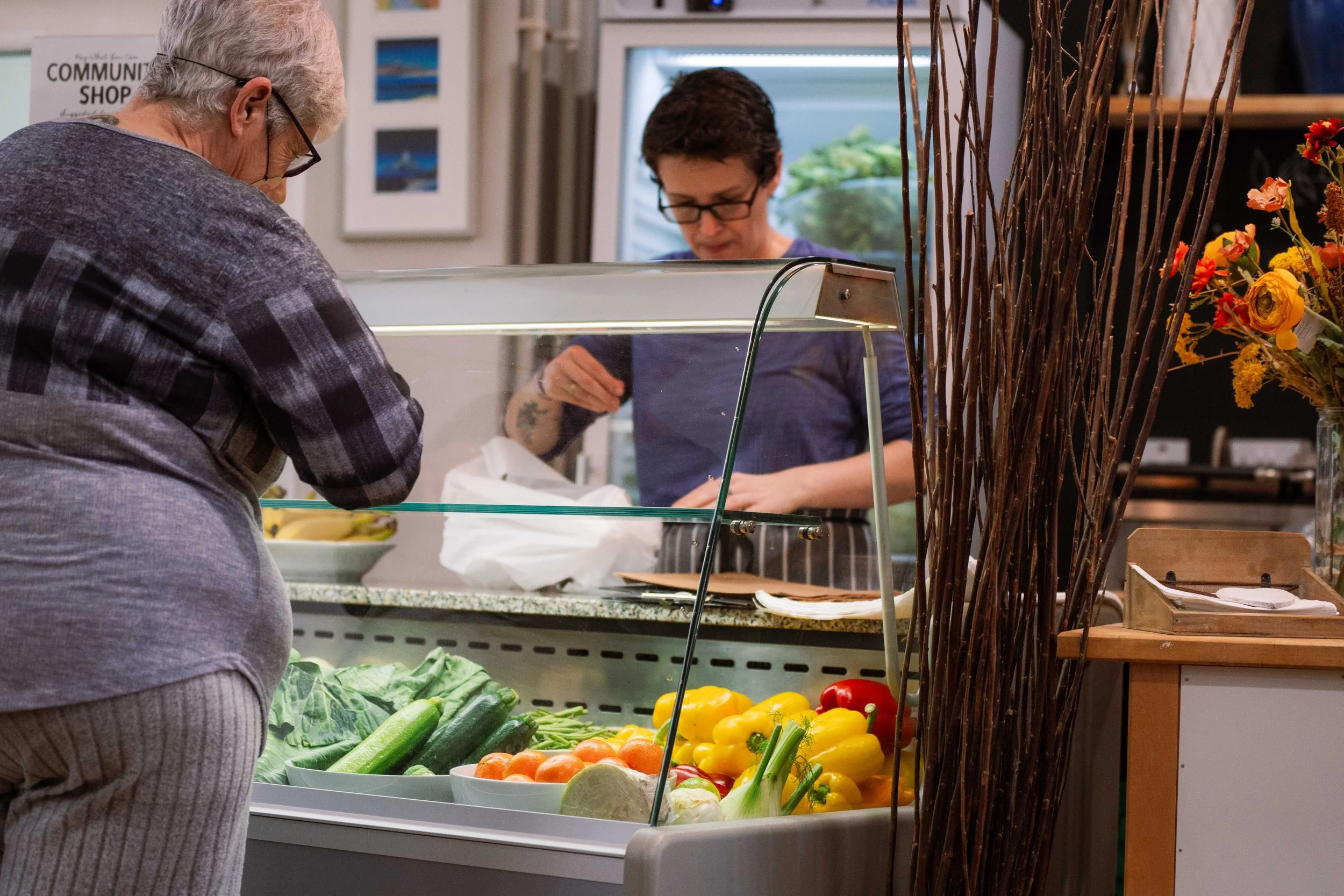 Two women at a grocery store or market, one selecting vegetables at a produce counter with cucumbers, yellow and red peppers, and other vegetables, while the other woman appears to be working behind the counter. Flowers and tall decorative sticks are also visible.