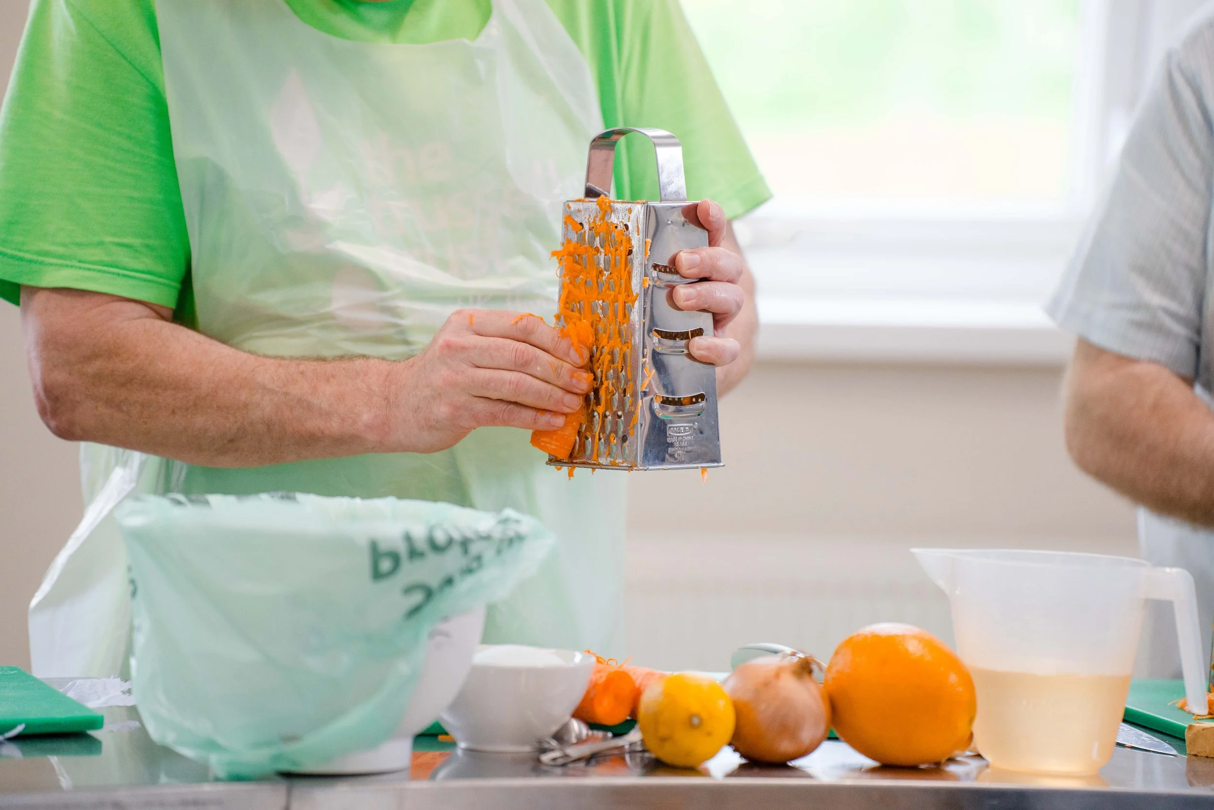 Person grating a carrot in a kitchen with various vegetables and ingredients on the counter.