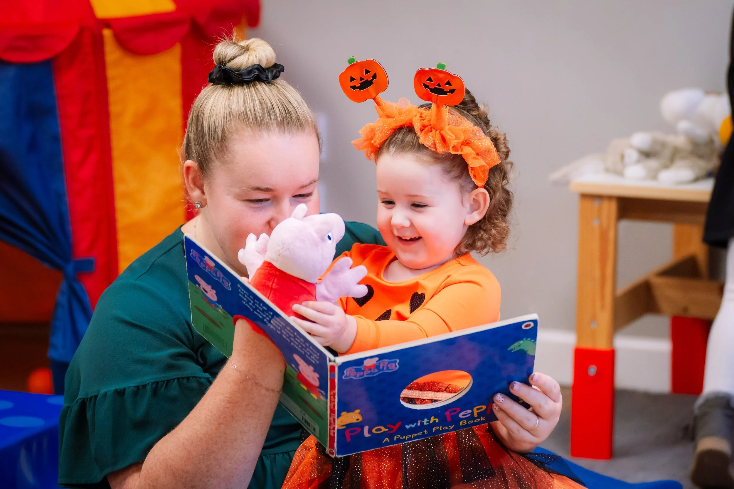 A woman and a young girl excitedly reading a puppet playbook together, with the girl wearing a Halloween-themed headband and bright orange costume.