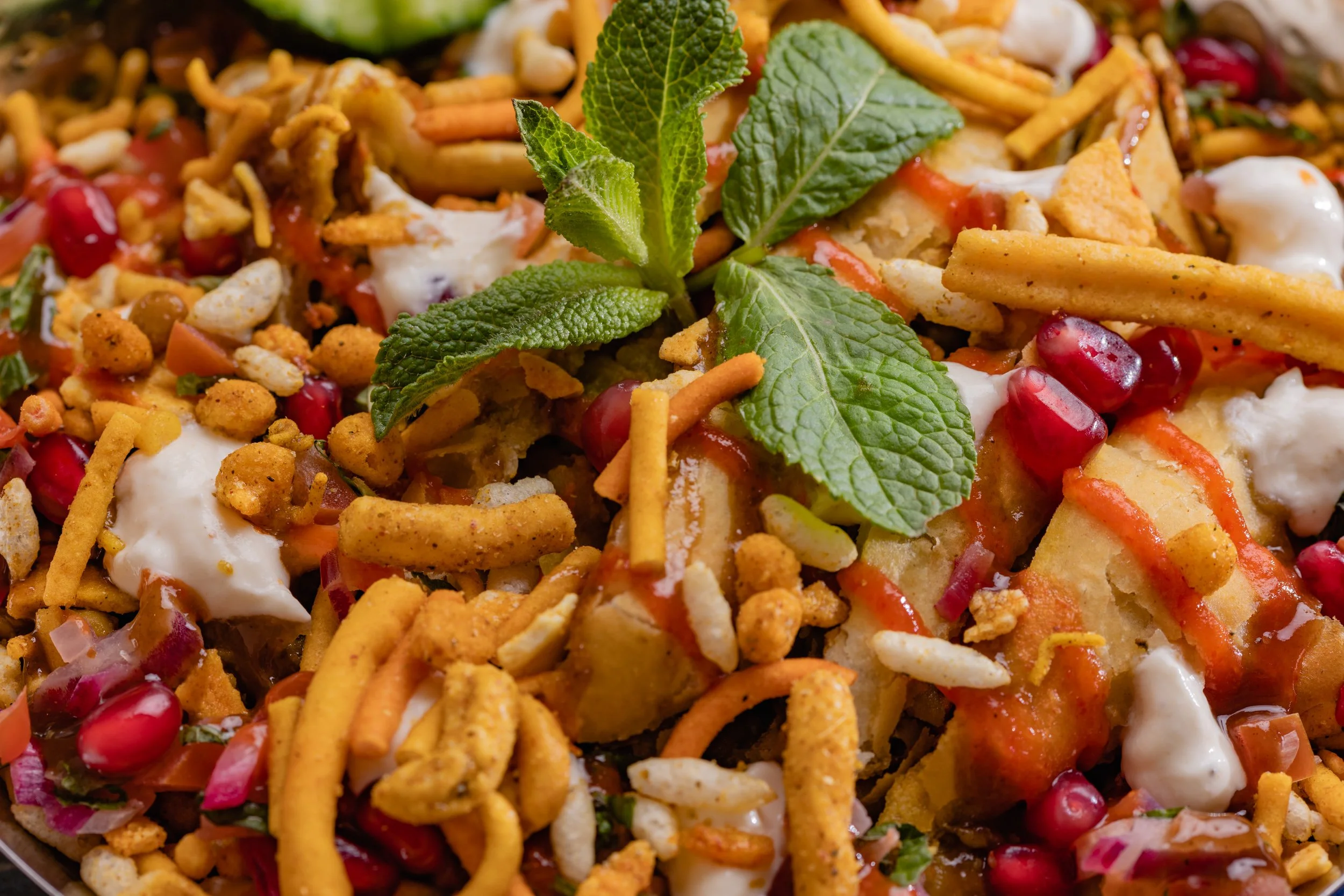 Close-up of a salad with pomegranate seeds, fresh mint leaves, crunchy noodles, chopped vegetables, and creamy dressing.