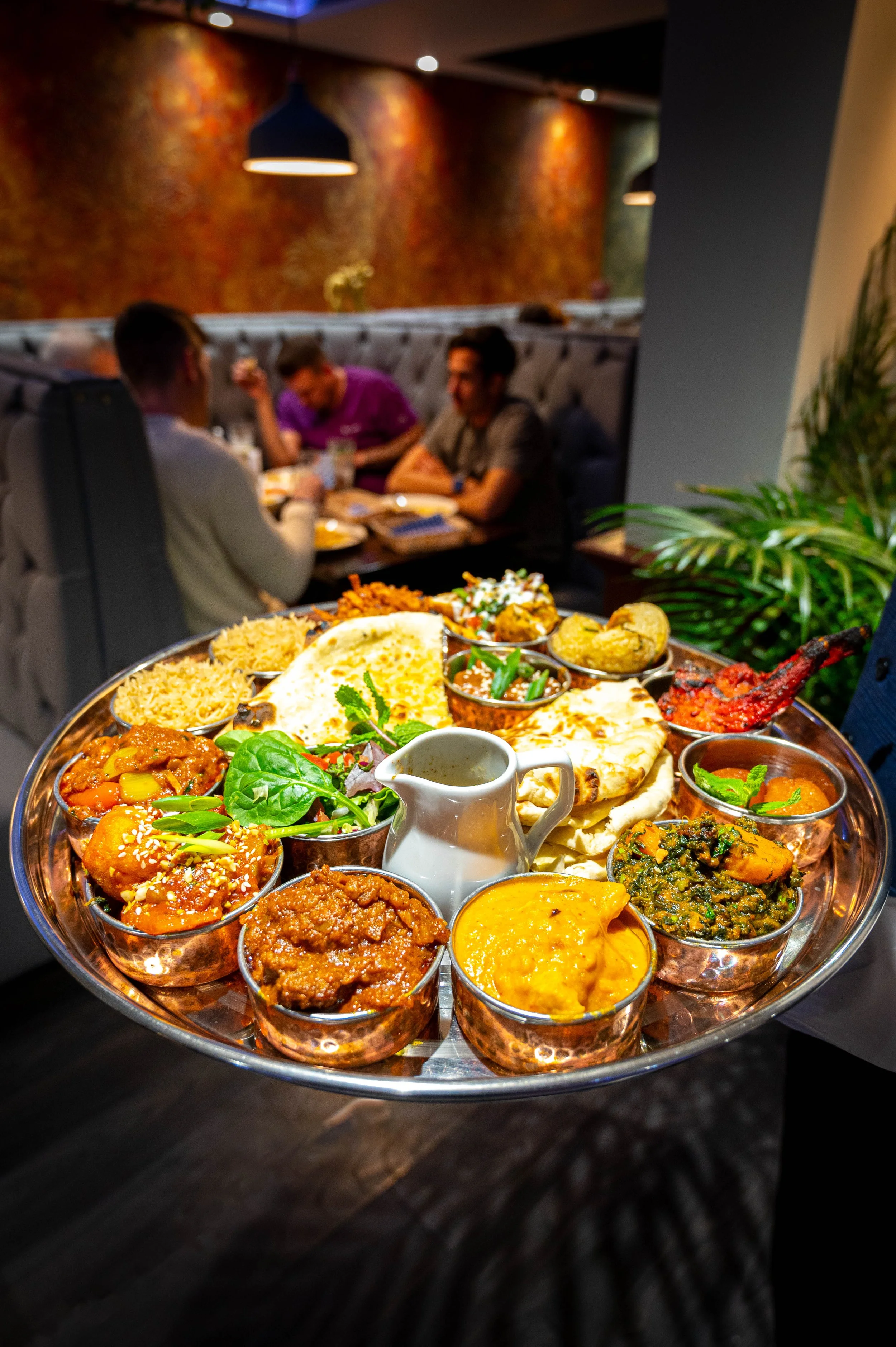 A large tray of Indian food with various curries, rice, naan bread, and side dishes on a dark table in a restaurant with three people dining in the background.