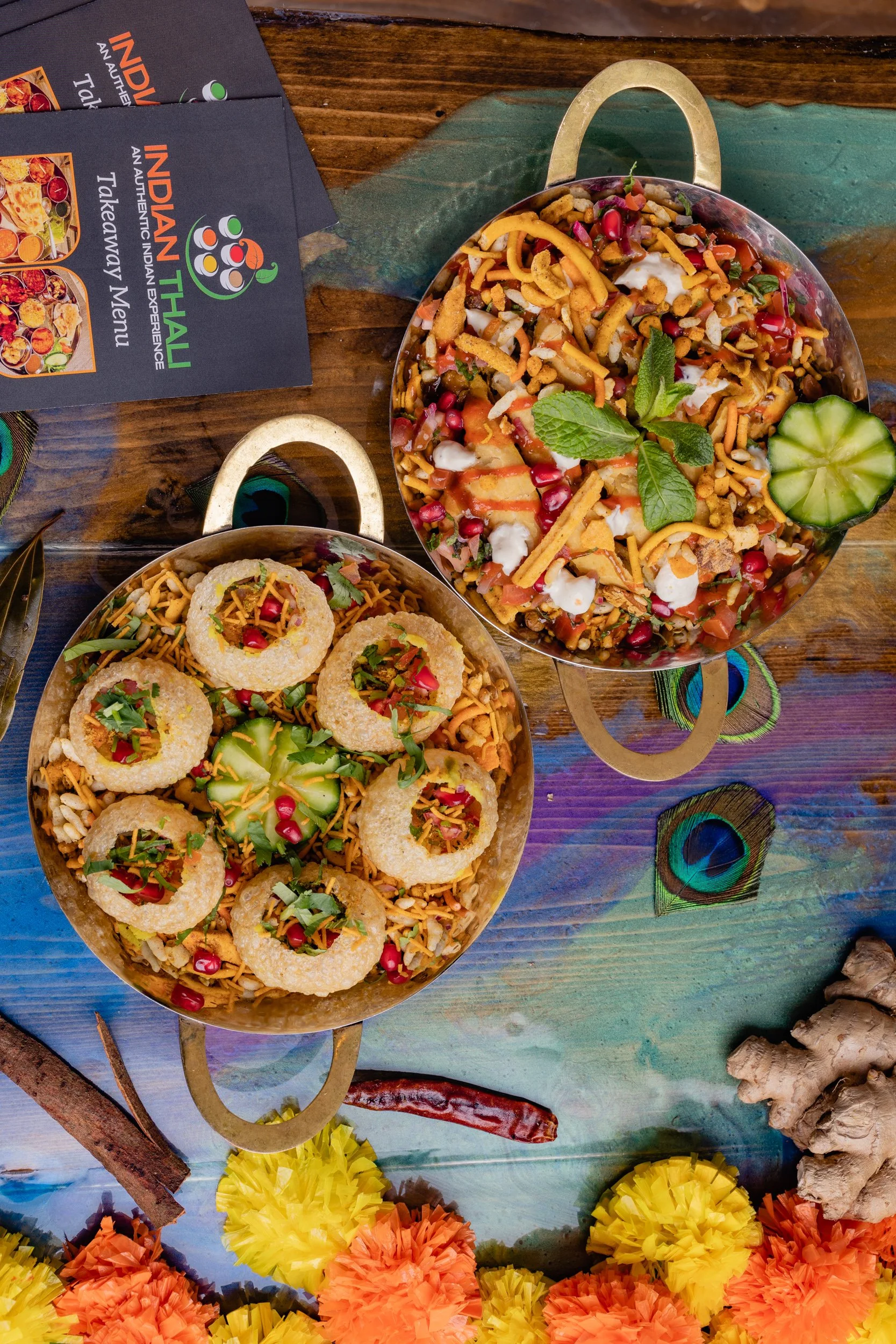 Two metal bowls filled with Indian snacks, garnished with herbs, placed on a colorful wooden table with spices, flowers, and Indian restaurant brochures nearby.