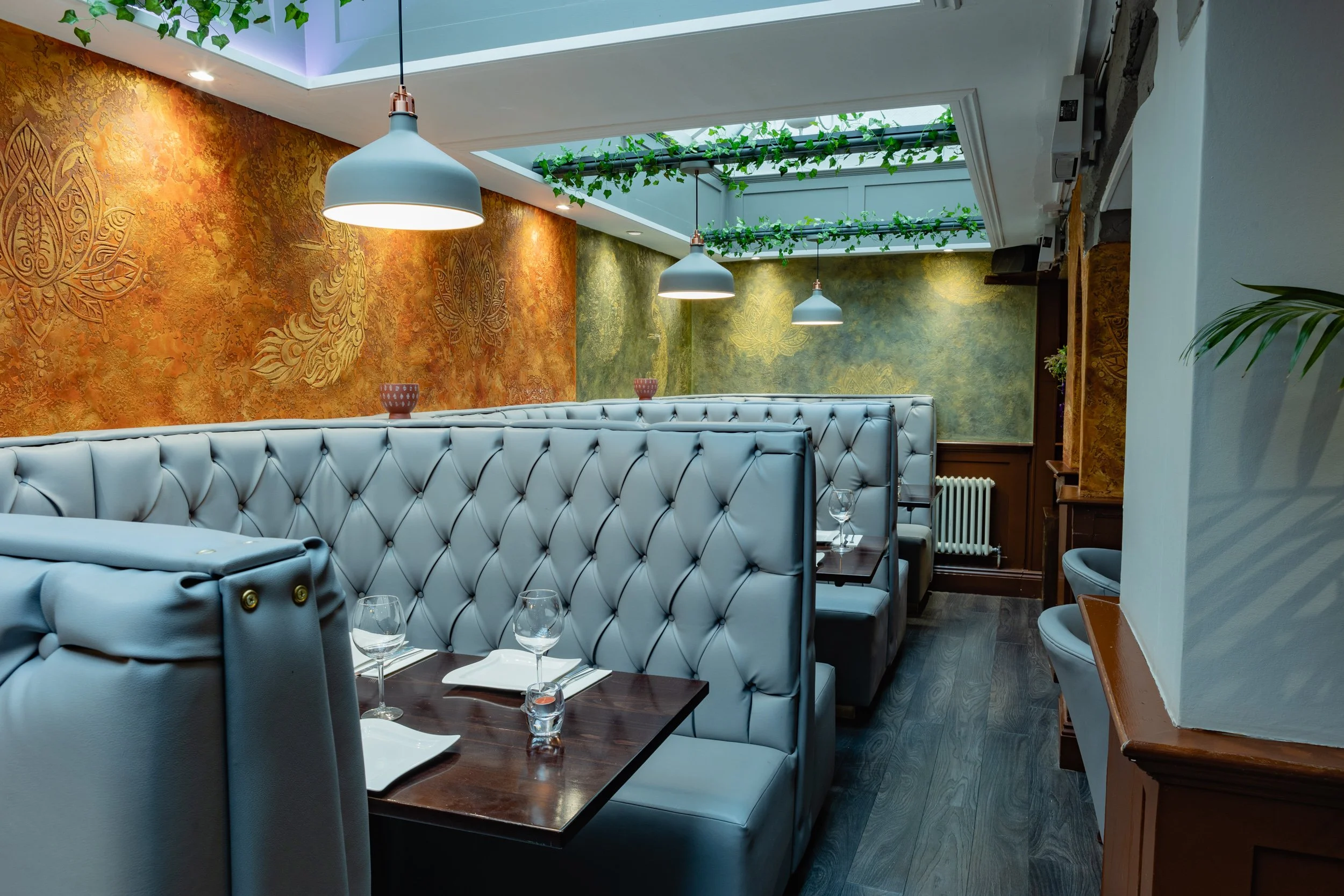 A cozy restaurant booth with high white leather tufted seats, set with wine glasses, napkins, and small bowls on dark wooden tables. The background features decorative gold and green textured walls and a ceiling with natural light from a skylight fil