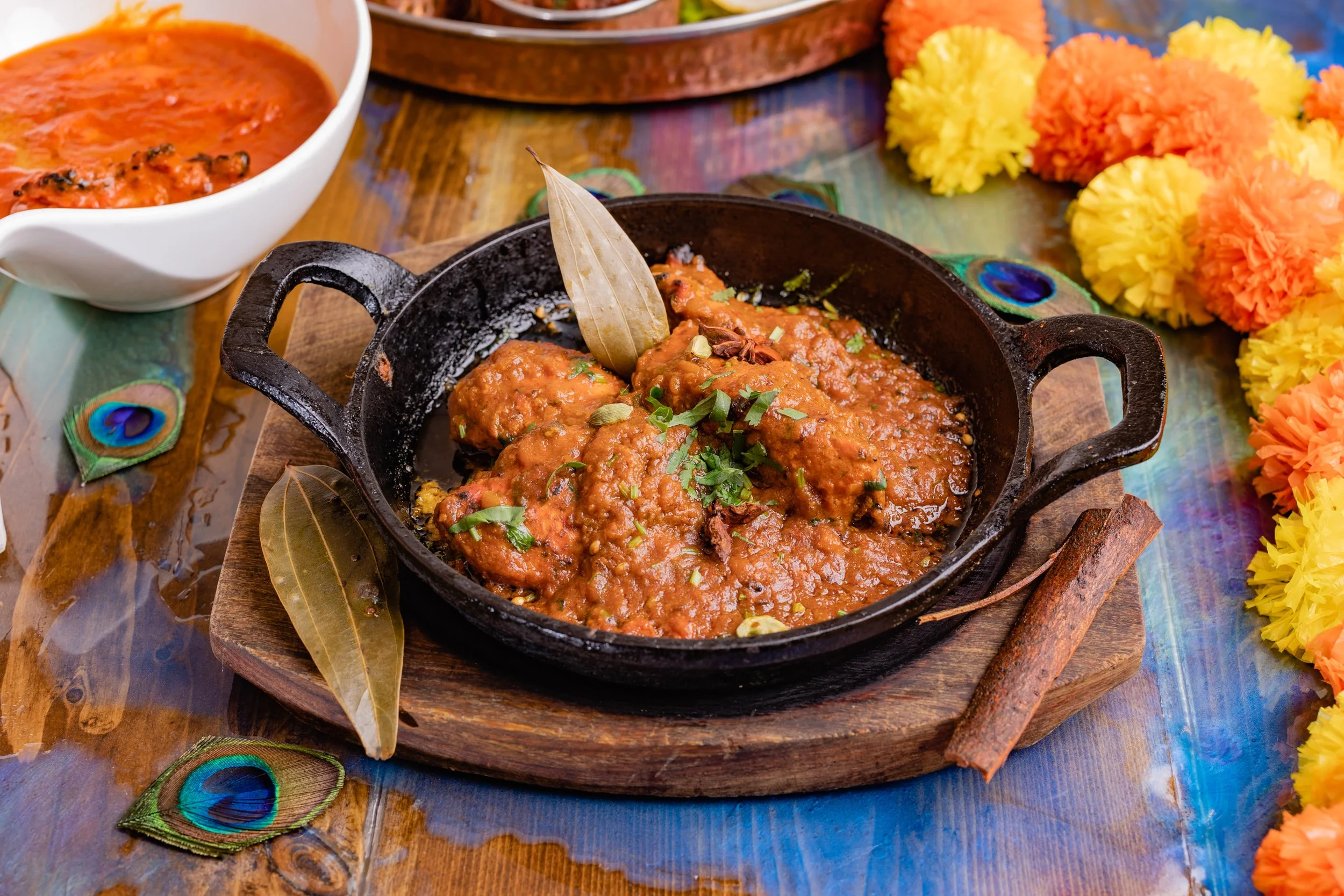 A serving of Indian chicken curry garnished with cilantro in a black cast iron dish on a wooden board, with a bay leaf, cinnamon stick, and decorative peacock feathers on a colorful table with marigold flowers and a bowl of tomato-based sauce.