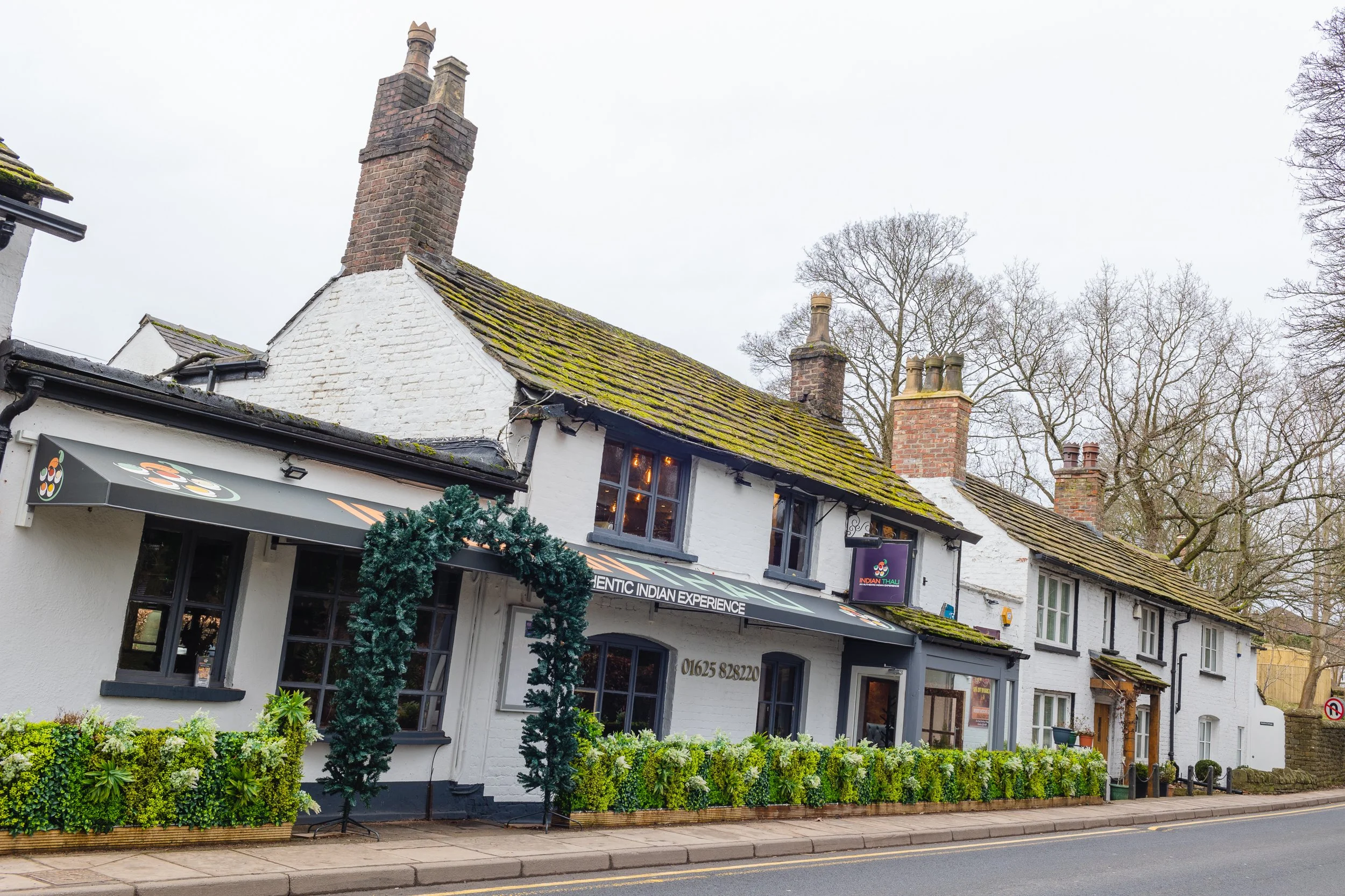 White building with mossy roof and black trim, decorated with greenery, houses a restaurant called Indian Thali, located on a street with trees in the background.