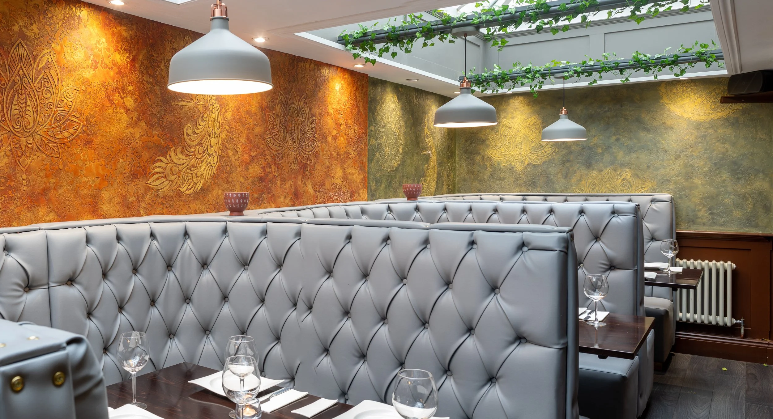 Upscale restaurant booths with tufted white leather seats, dark wood tables set with wine glasses, in front of brightly colored textured walls with floral and peacock designs, under hanging white pendant lights, with a skylight and greenery above.