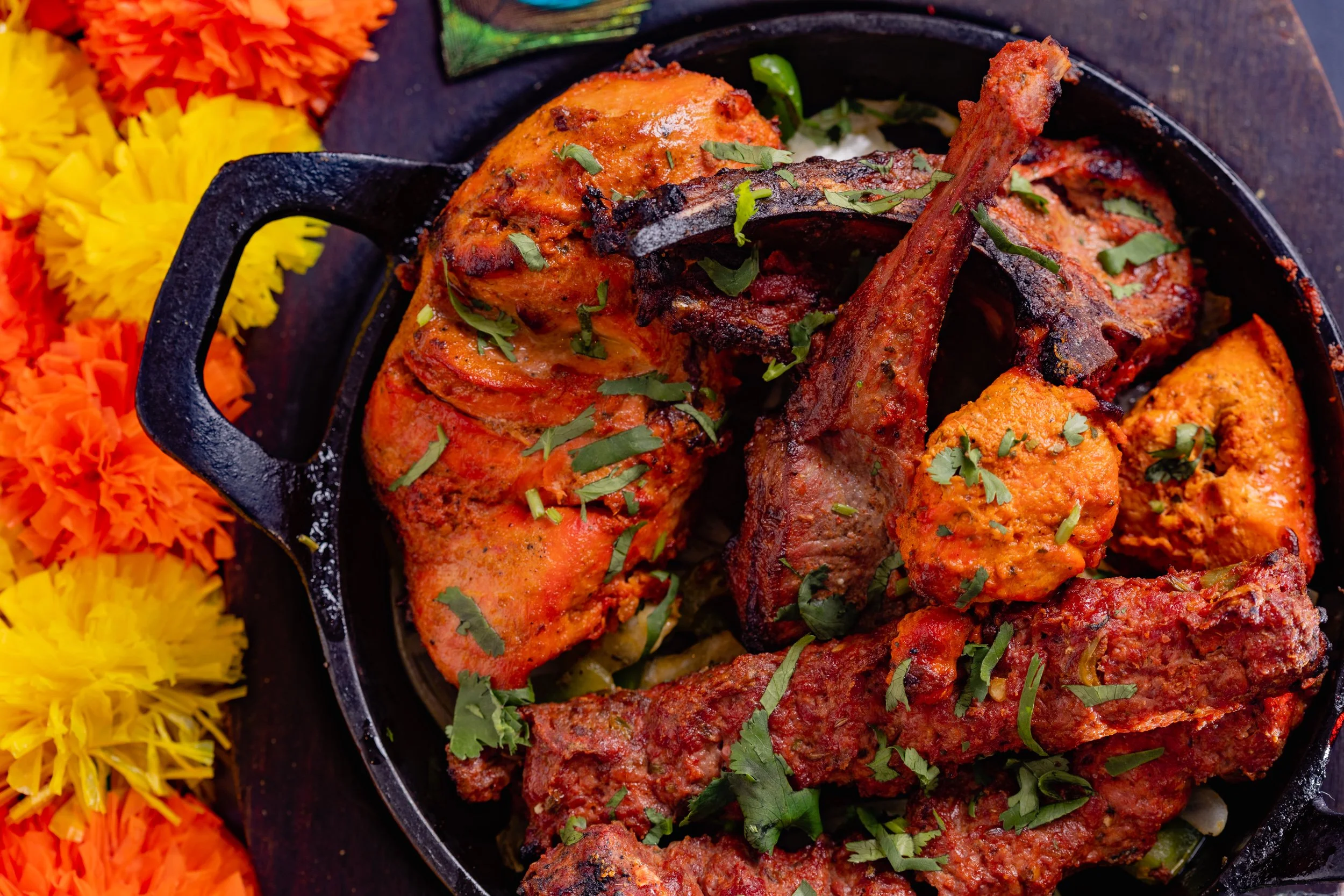 Assorted Indian grilled meats, including chicken, lamb chops, and beef, garnished with chopped cilantro and served in a black cast iron skillet, with colorful Indian marigold flowers decorate around the skillet.
