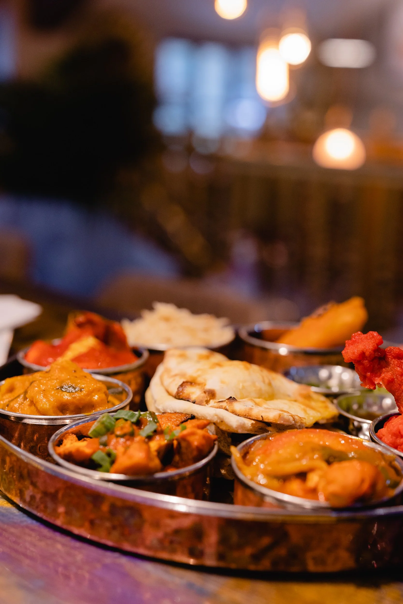 A round silver tray with various Indian dishes, including curries, naan bread, and chutneys, set on a wooden table in a dimly lit restaurant.