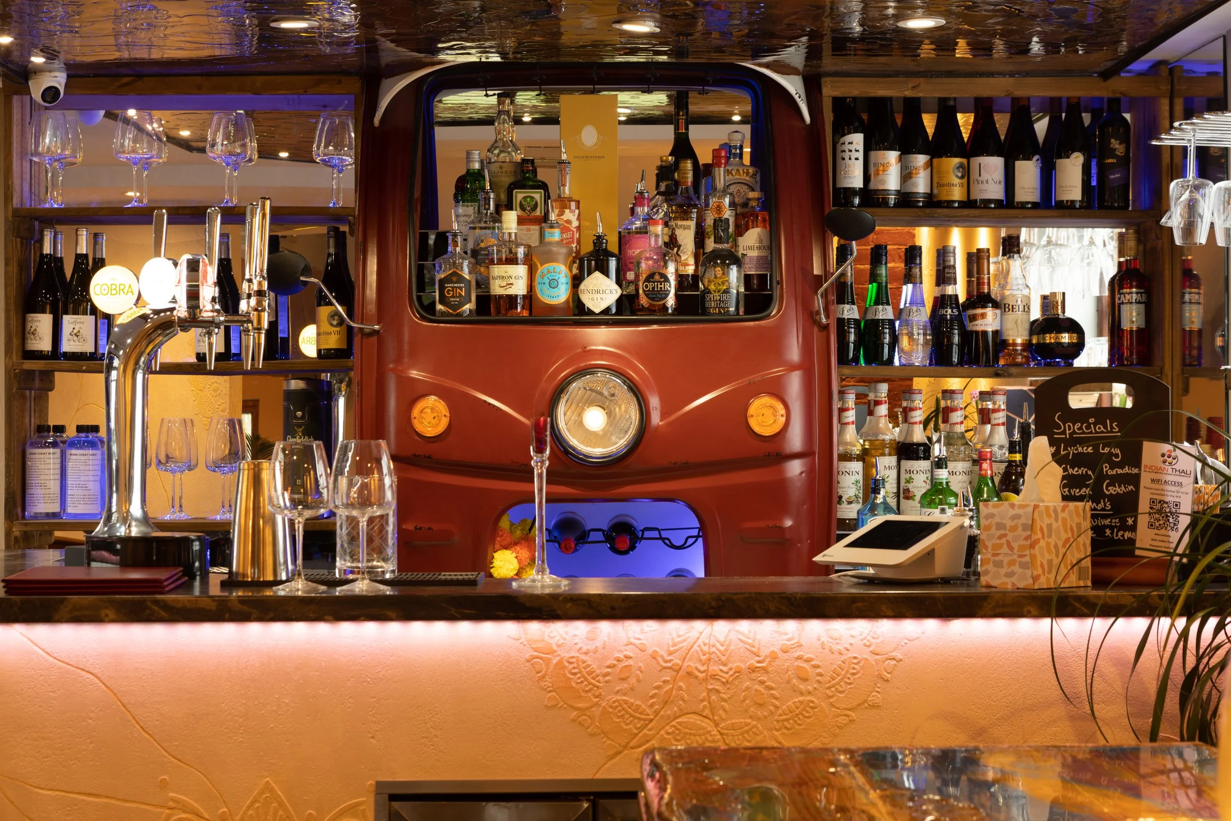 Bar counter with a red trailer front as shelving behind it, stocked with various bottles of liquor and wine, with glassware and bar equipment. The counter has glasses, a small tap, and a digital payment device, with warm lighting and decorative eleme