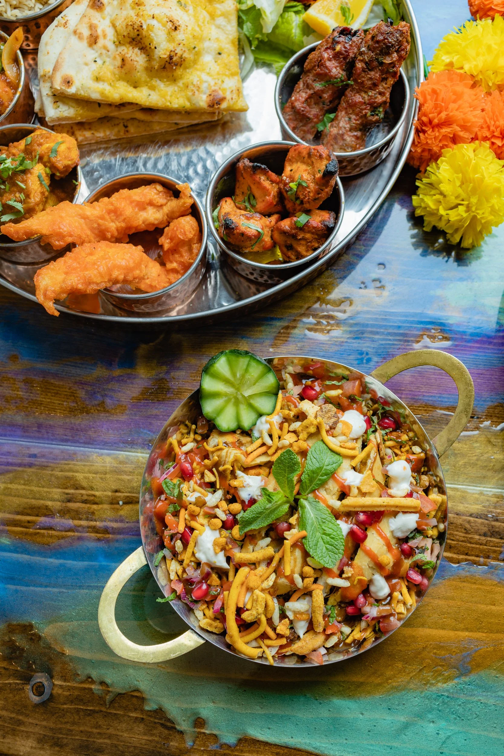 A colorful Indian meal featuring a bowl of bhel puri topped with cucumber slices and mint leaves, a grilled vegetable chaat, and a platter of assorted Indian snacks, including naan, samosas, and spiced meats, with yellow and orange marigold flowers o