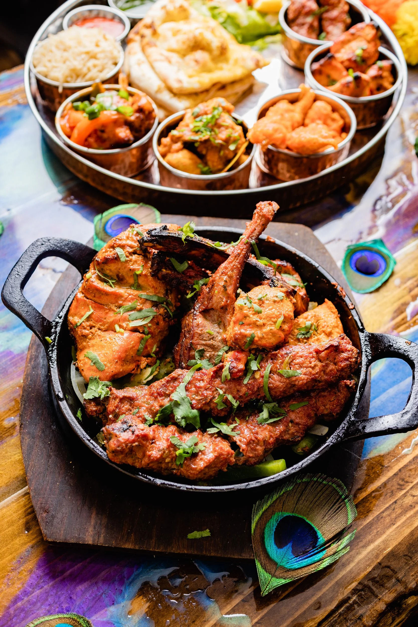 Close-up of a cast iron skillet with cooked pieces of chicken in sauce, garnished with cilantro, on a wooden board. In the background, an Indian meal with various curry dishes, naan bread, rice, and vegetable dishes on a circular tray.