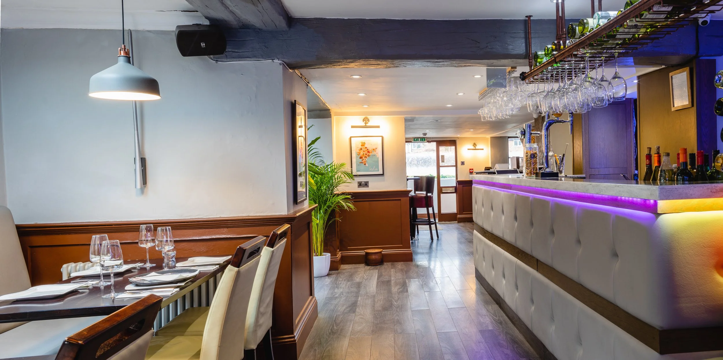 Interior of a restaurant or bar with a white bar counter illuminated with purple and yellow lighting, glasses hanging upside down, and a variety of bottles behind the bar. In the dining area, there are tables set with glasses, napkins, and cutlery, w