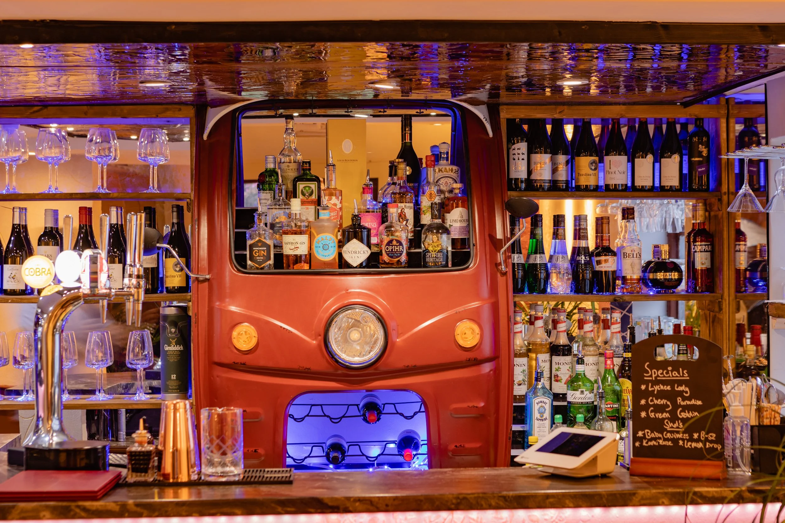 Bar with a vintage red truck front as the display, stocked with liquor bottles, glassware, and a chalkboard menu, in a warm, inviting setting.