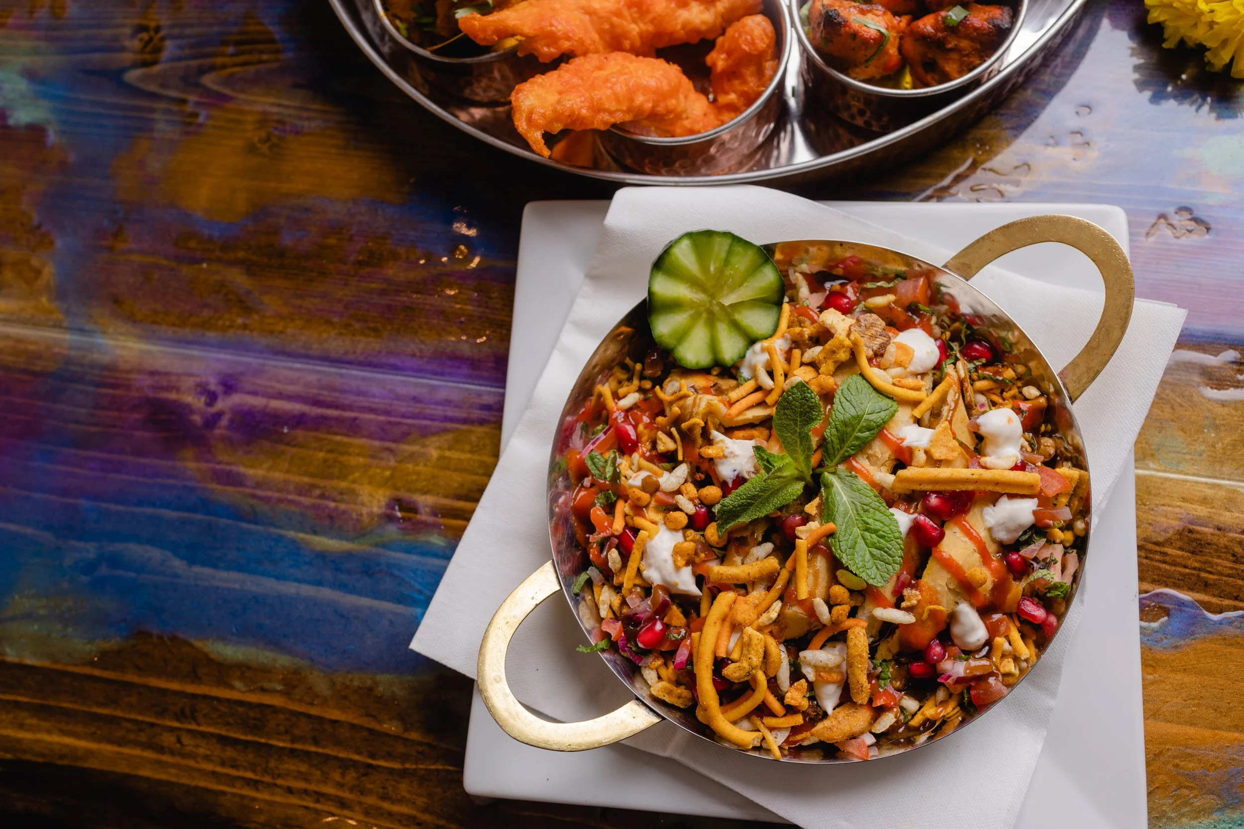 A bowl of Indian chaat garnished with mint leaves and a cucumber slice, with fried snacks and chickpeas in the background.