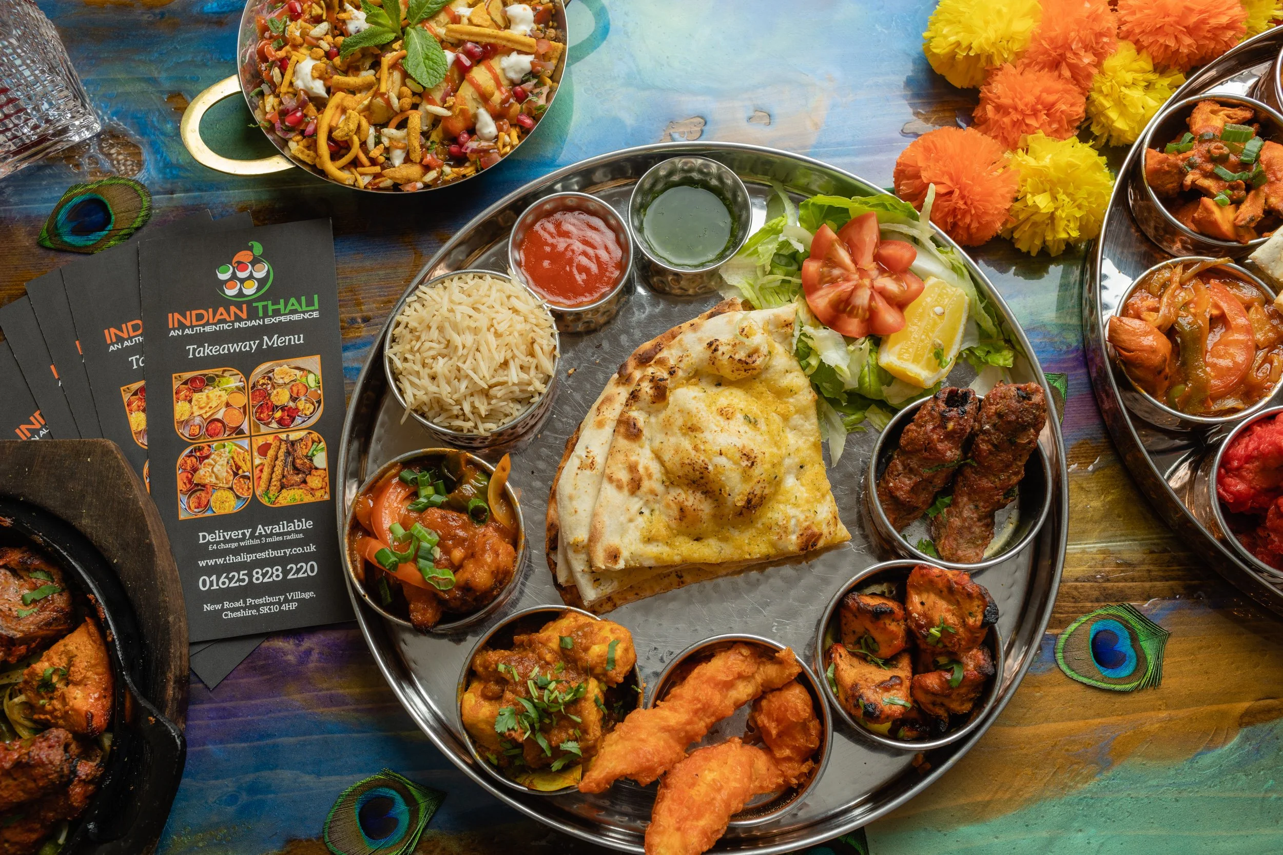 An Indian meal featuring naan bread, rice, various curries, and side dishes served on a large silver platter, surrounded by small bowls of chutneys and fresh salad, with a colorful table and decorative flowers.