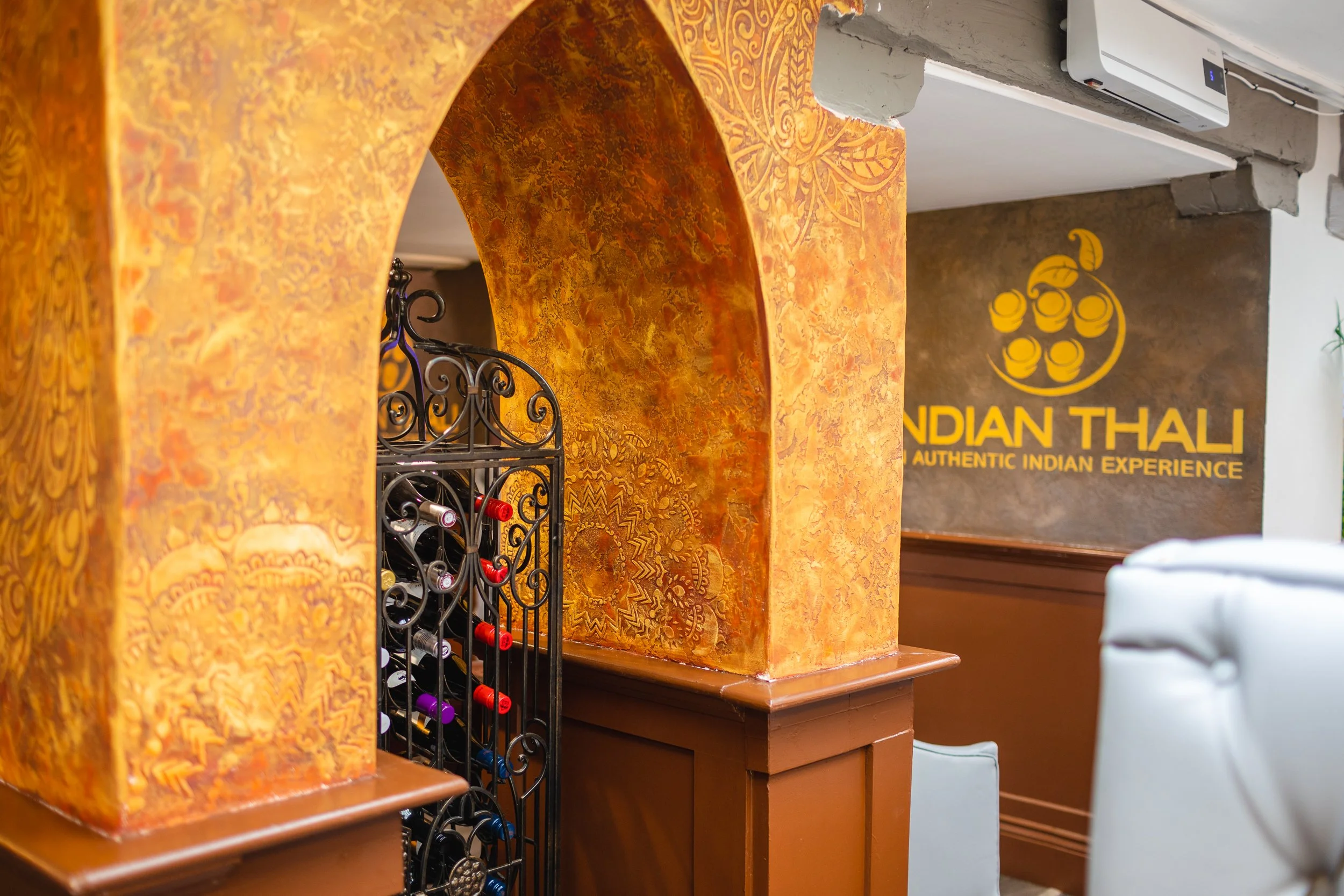 Interior of a restaurant with orange textured walls, a black wrought iron wine rack, and a yellow sign with a logo and text that reads 'INDIAN THALI - AUTHENTIC INDIAN EXPERIENCE' in the background.