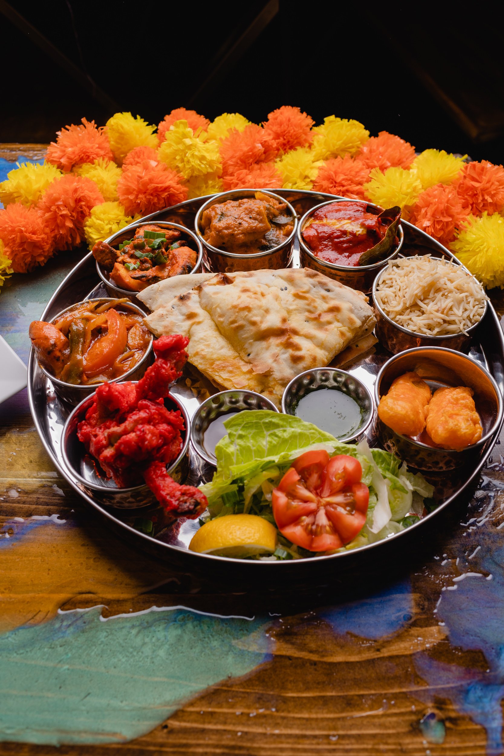 An Indian Thali meal on a metal platter with various curries, rice, naan, salad, and lemon slice, surrounded by orange and yellow marigold flowers.