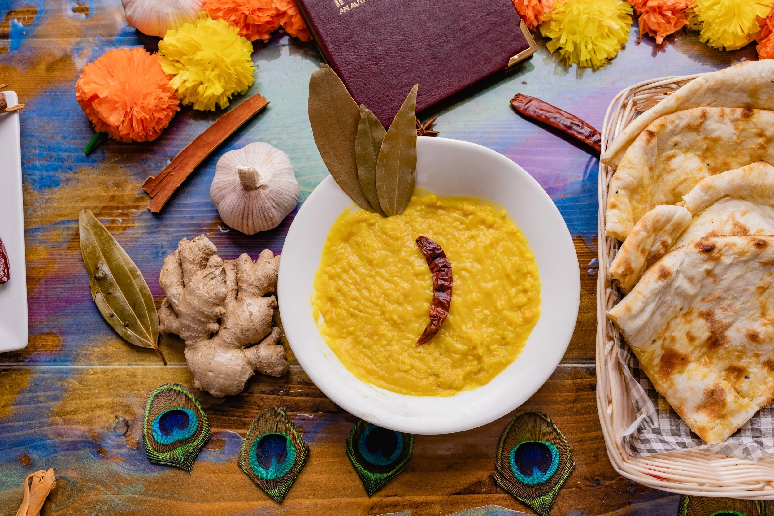 A white bowl of yellow lentil soup garnished with a dried red chili on top and two bay leaves placed inside it. Surrounding the bowl are ginger, garlic, a dried chili, a marigold flower, a maroon book, and a basket of cooked flatbread. Peacock feathe
