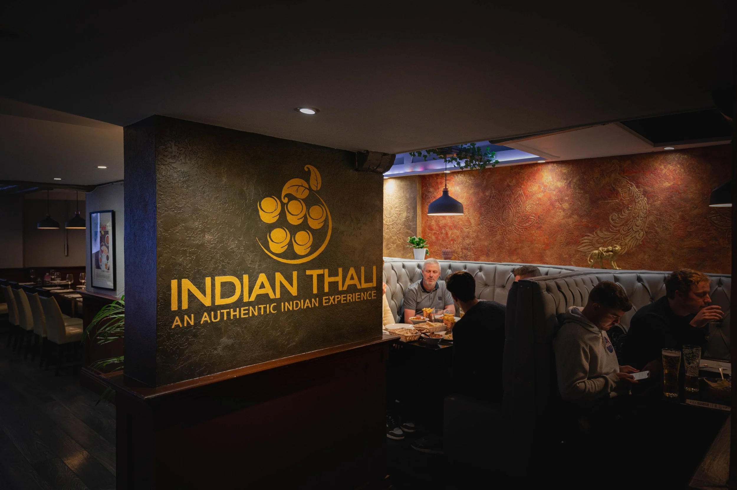 Interior of an Indian restaurant with a sign that reads 'Indian Thali - An Authentic Indian Experience.' Several people are dining at tables, with food and drinks visible.