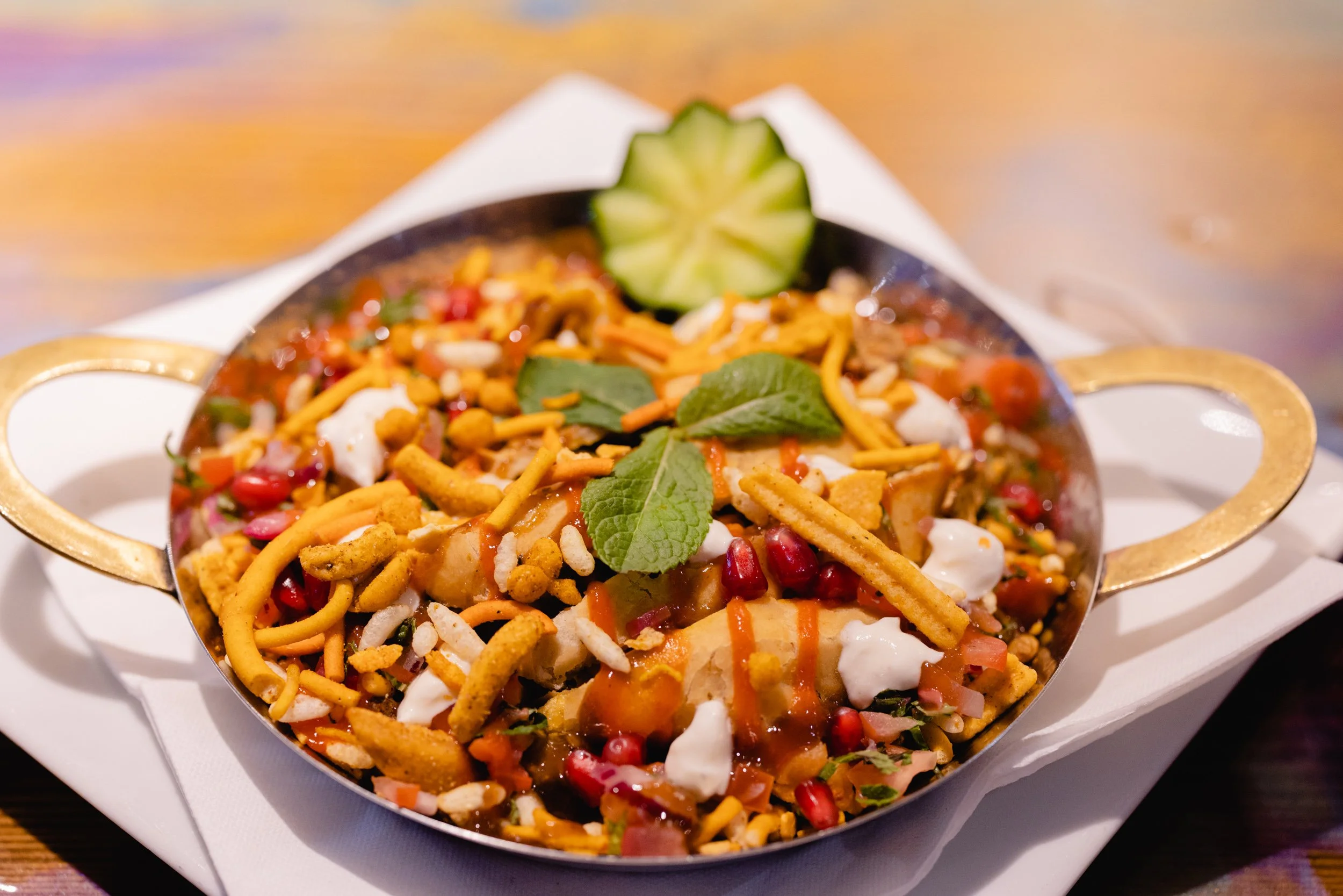 A colorful Indian street food dish served in a small brass bowl, garnished with fresh mint leaves and a cucumber slice, featuring noodles, pomegranate seeds, vegetables, and spices.