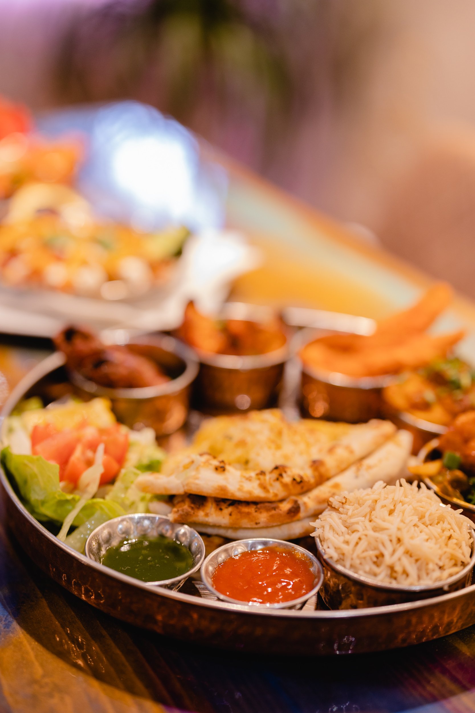 A large round tray of Indian food including naan bread, rice, various curries, sauces, and a salad with tomatoes and lettuce.