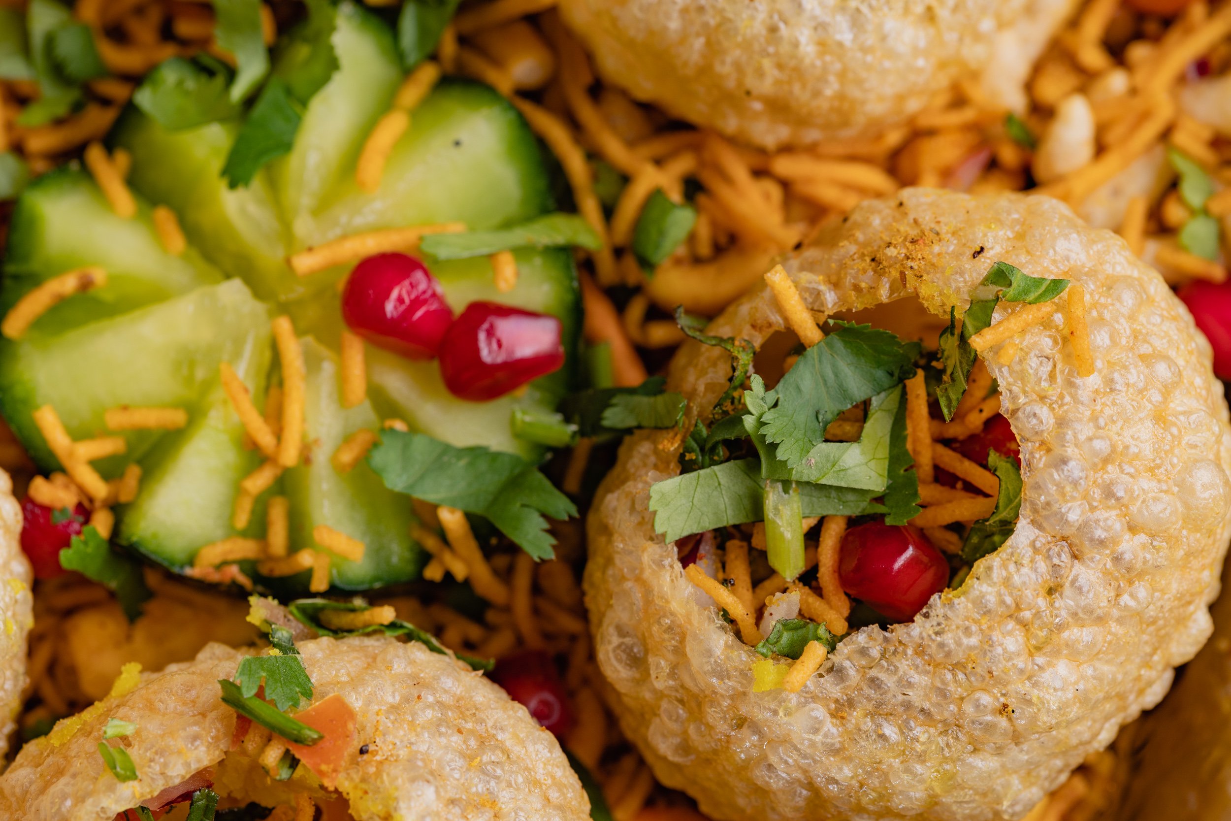 Close-up of Indian street food dish showing pani puri shells filled with chickpeas, topped with cilantro, with sliced cucumbers, pomegranate seeds, and sev in the background.