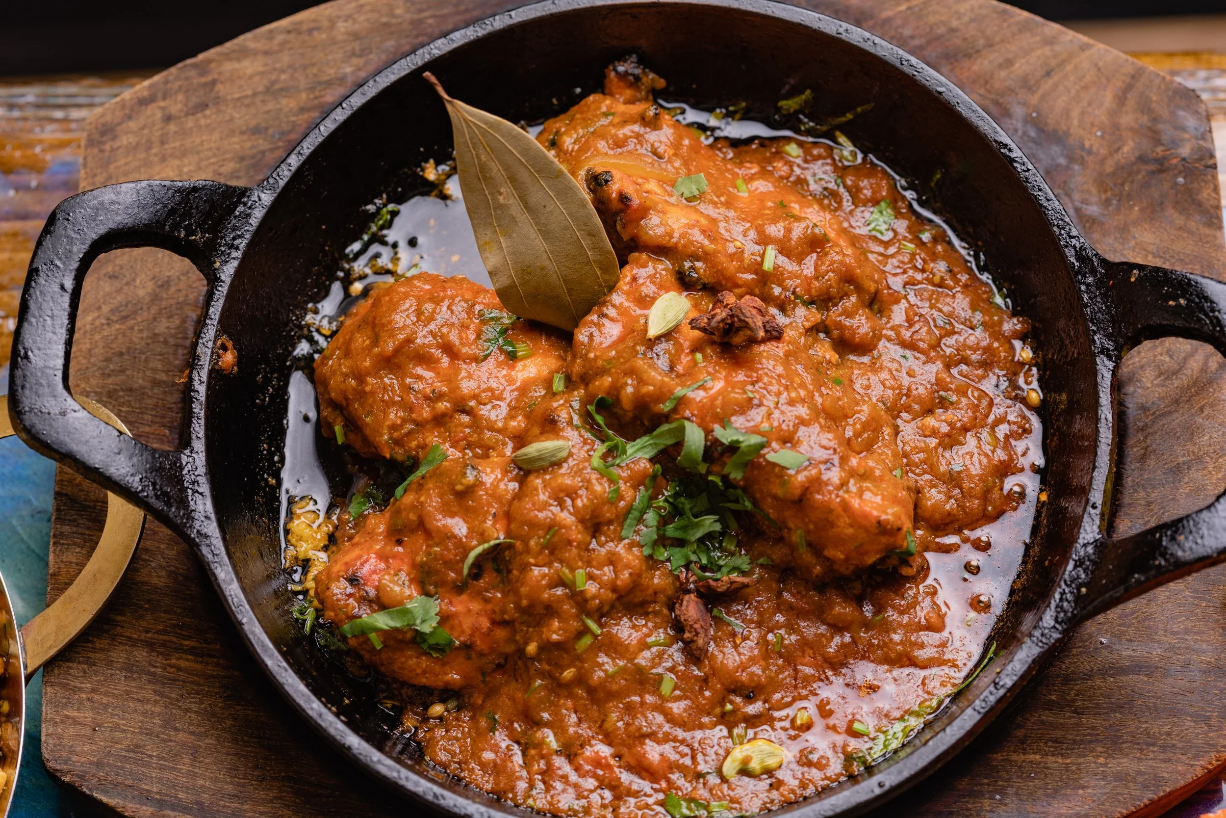 Cooked fish in a spicy tomato-based sauce garnished with coriander leaves, bay leaf, and cardamom pods in a black cast iron skillet on a wooden board