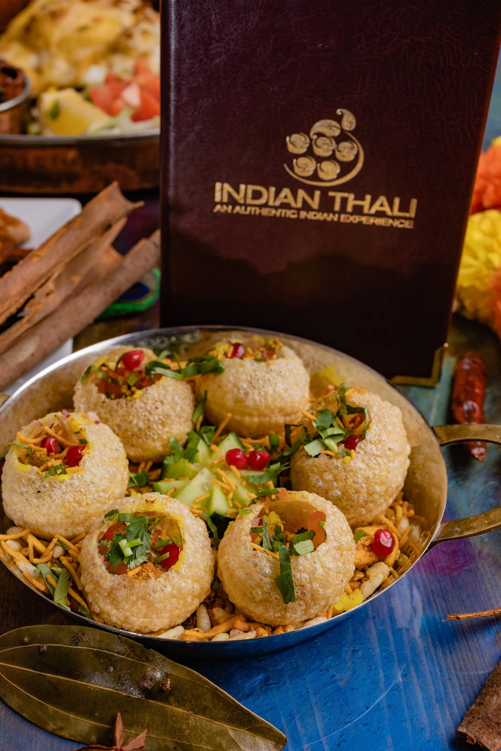 Plate of Indian-style stuffed bread balls garnished with herbs and pomegranate seeds, with a menu/menu cover labeled 'Indian Thali' in the background.