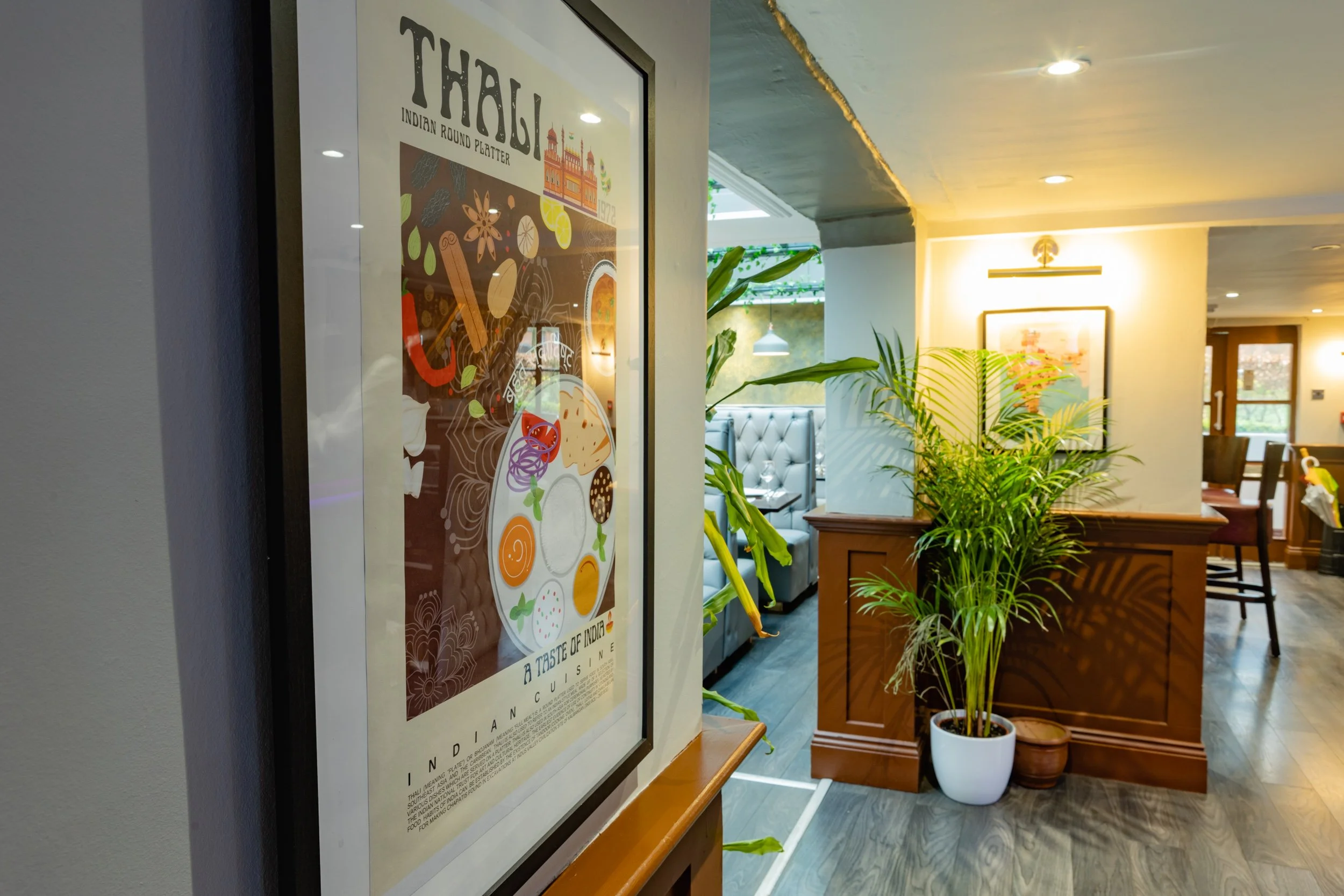 Interior of a restaurant with a framed poster of Indian cuisine on the wall, potted plants, a gray booth, and wooden furniture.