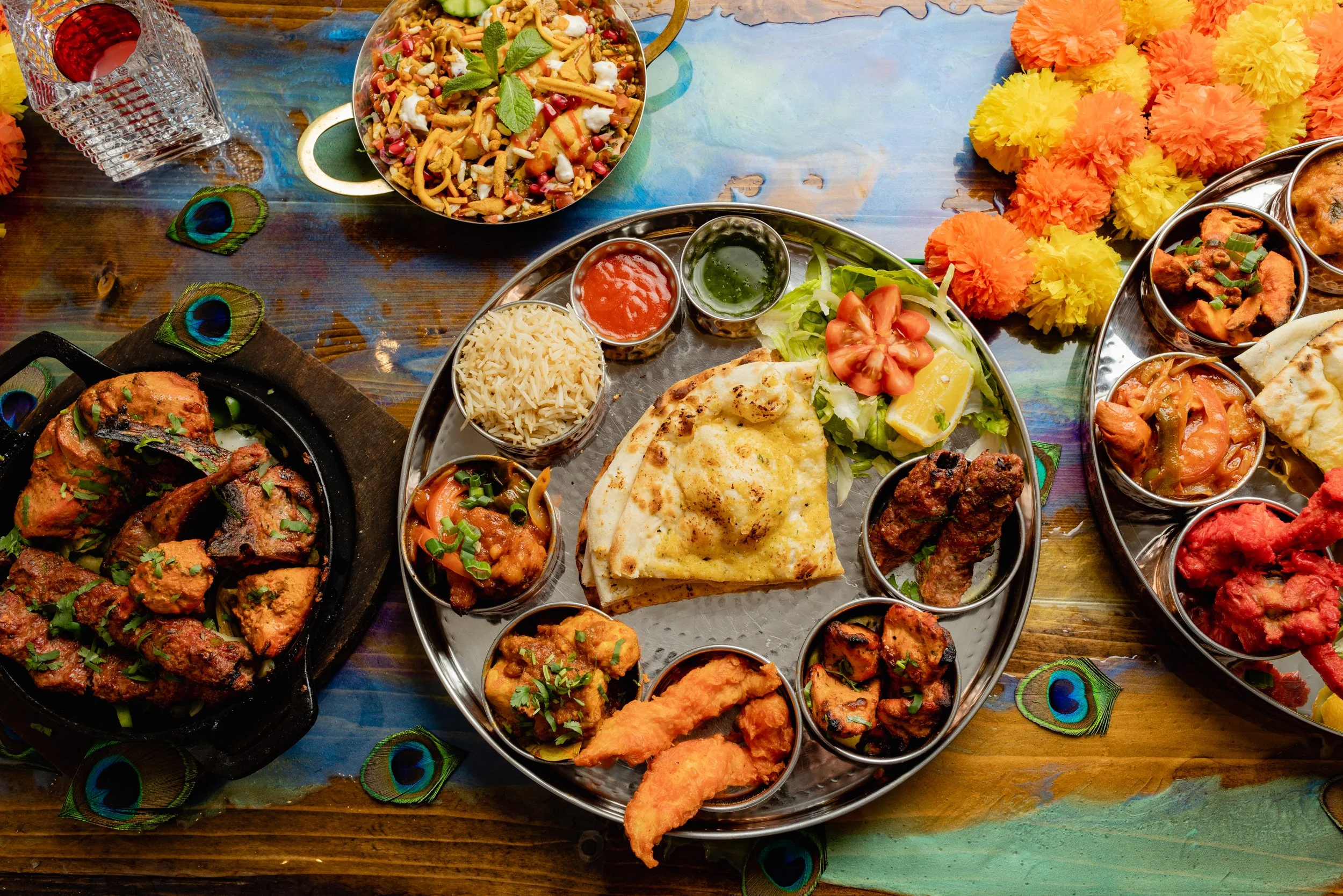 A table with Indian dishes including naan, rice, chicken curry, vegetable curries, and side salads, decorated with colorful marigold flowers and peacock feathers.
