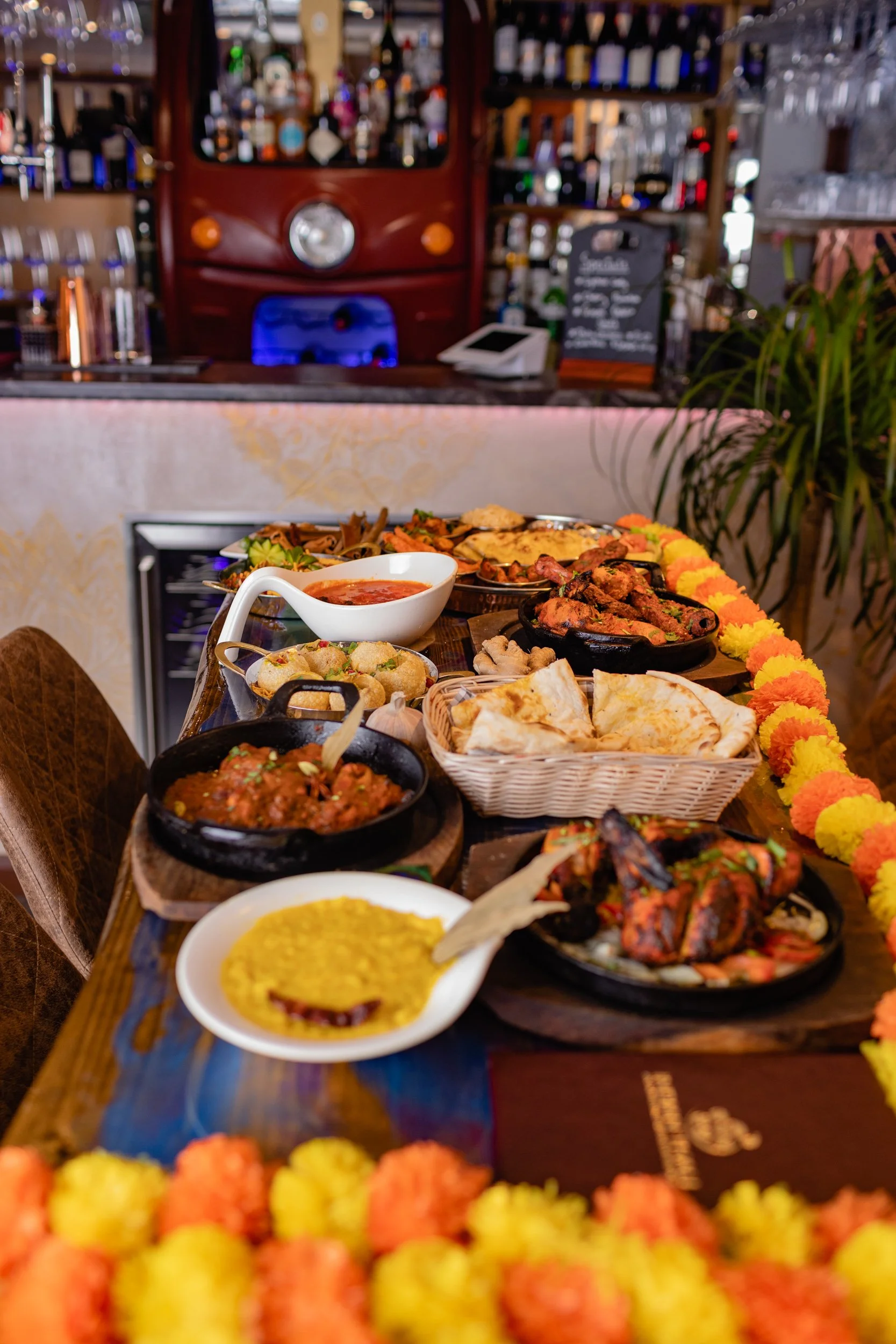 A table filled with Indian dishes including curry, naan bread, and various appetizers, decorated with marigold flower garlands in a restaurant.