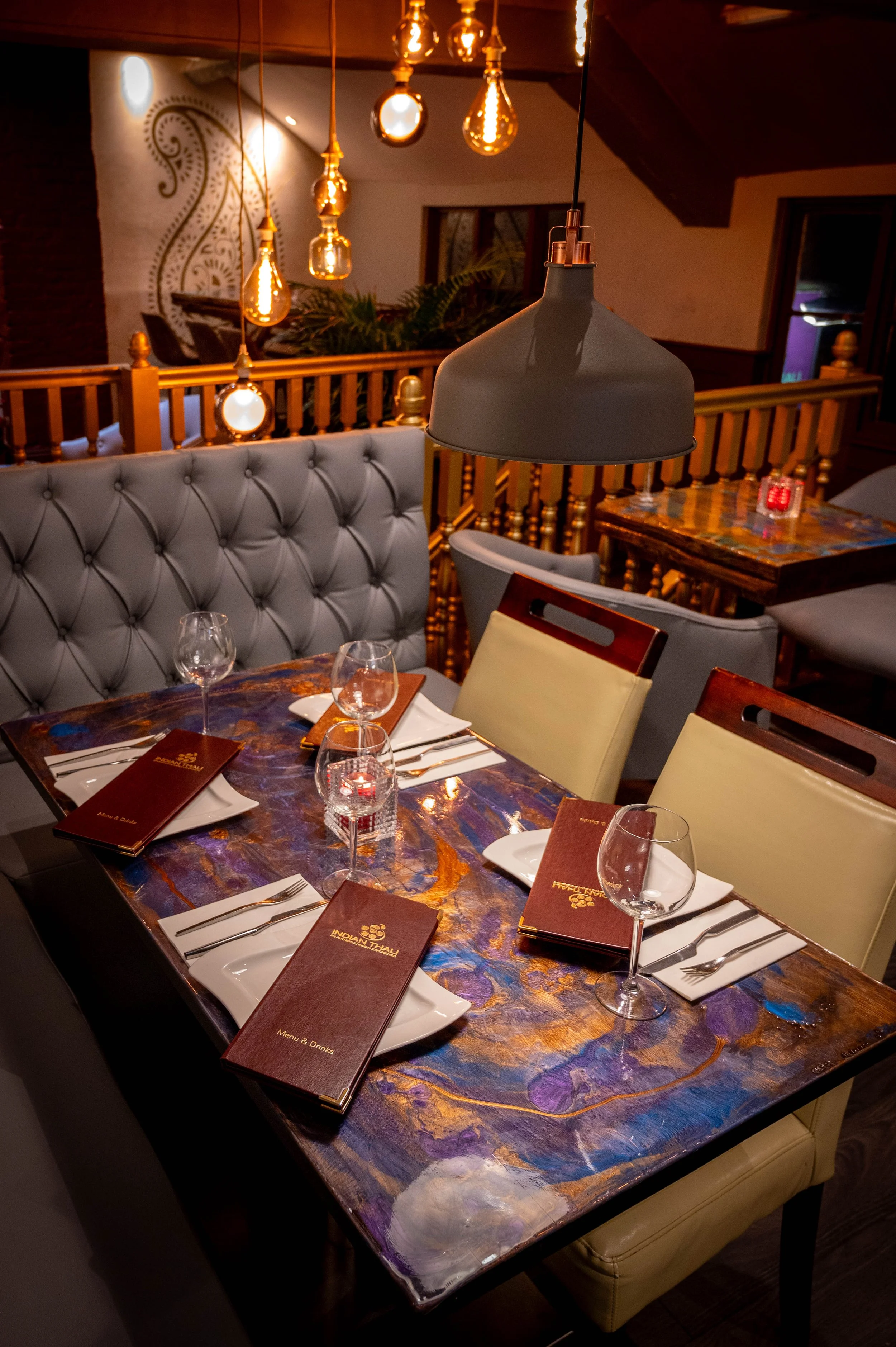 A restaurant dining table set with menus, wine glasses, and silverware, with a tufted leather cushioned bench, modern lighting fixtures, and warm ambient lighting in the background.