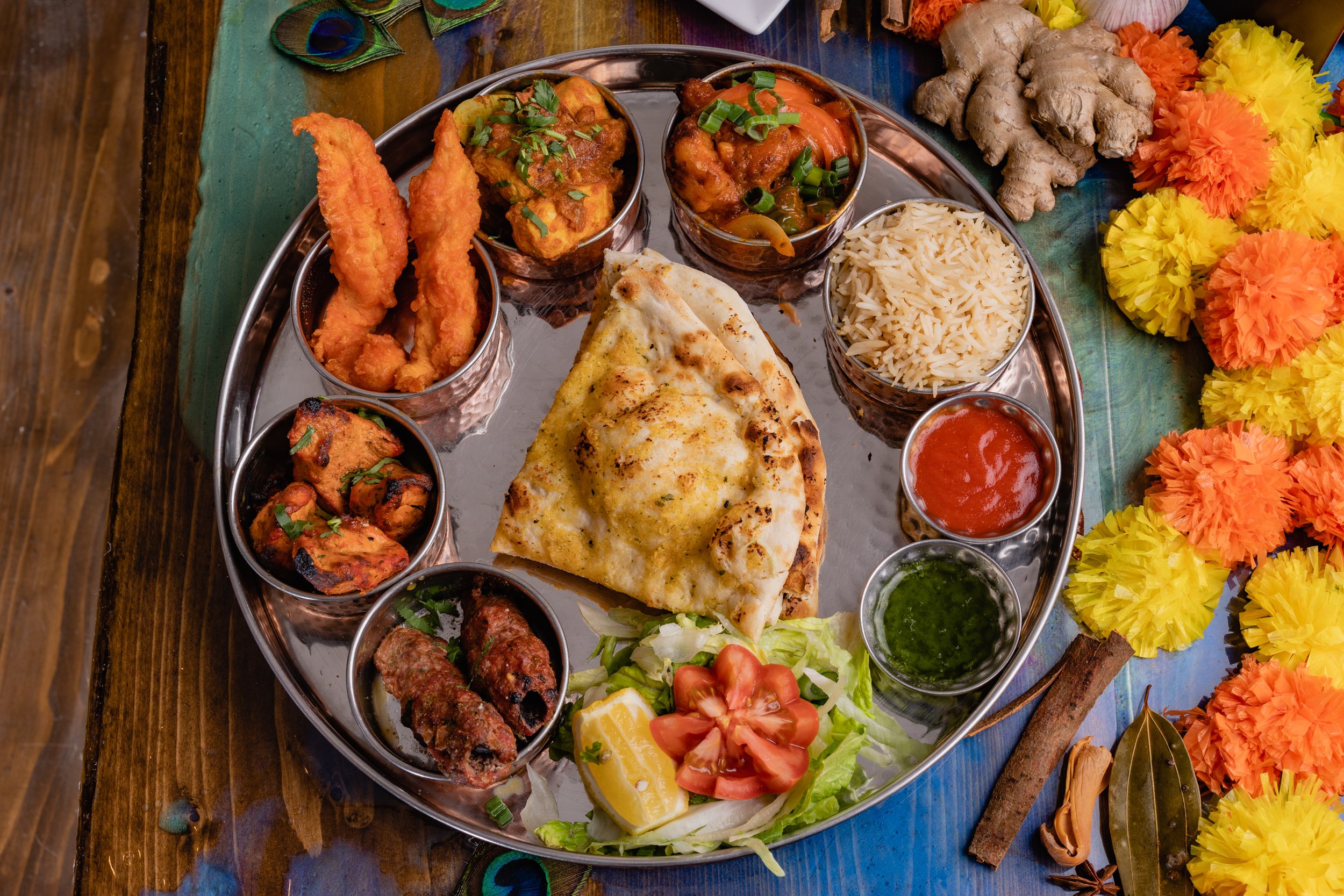 A traditional Indian meal served on a metal platter with various dishes including naan bread, rice, and assorted curries, garnished with salad, lemon, and herbs, surrounded by colorful decorations.