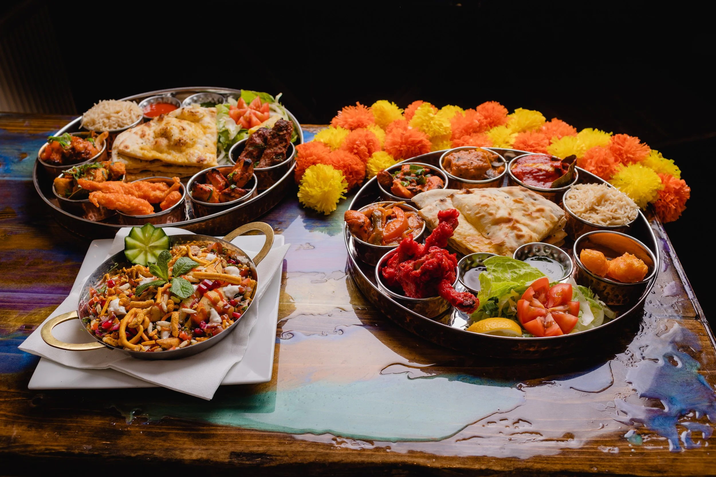 Two large round trays filled with Indian foods and a small dish of mixed snack mix on a colorful wooden table, surrounded by yellow and orange marigold flower decorations.