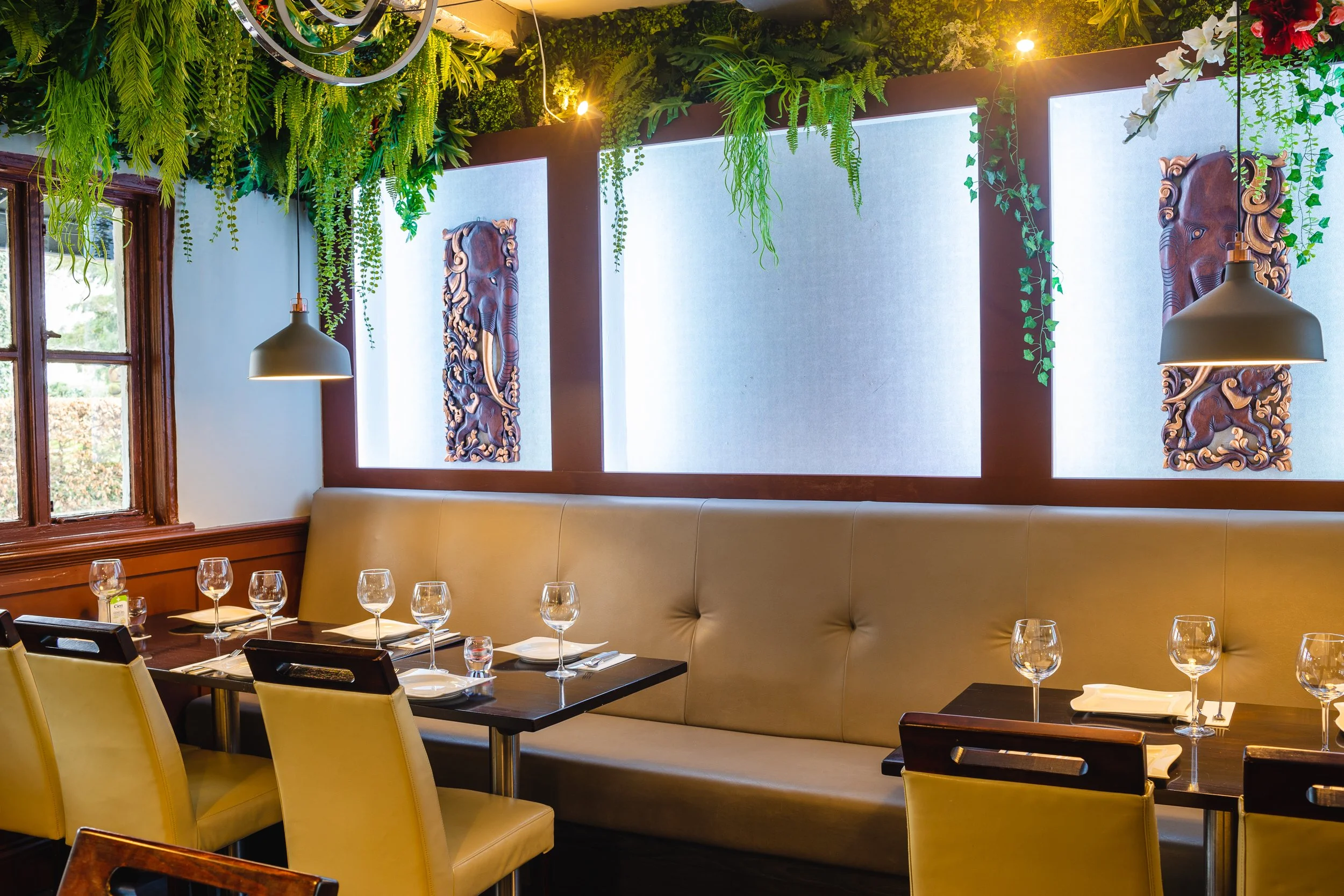 An empty restaurant with tan upholstered seating, dark wood tables with place settings, wine glasses, and white napkins. Decor includes green hanging plants and Asian-style wooden carvings of elephants on the wall, with large windows on the side.