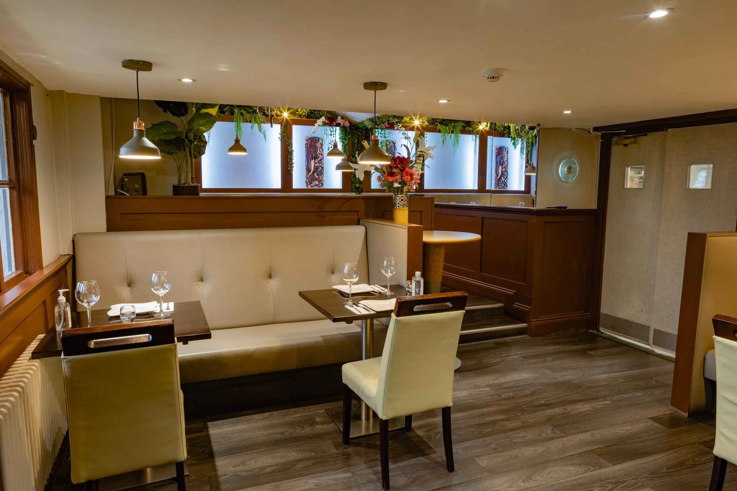 Empty restaurant booth with set tables, wine glasses, and a flower arrangement, featuring wooden floors and decor with plants and hanging lights.