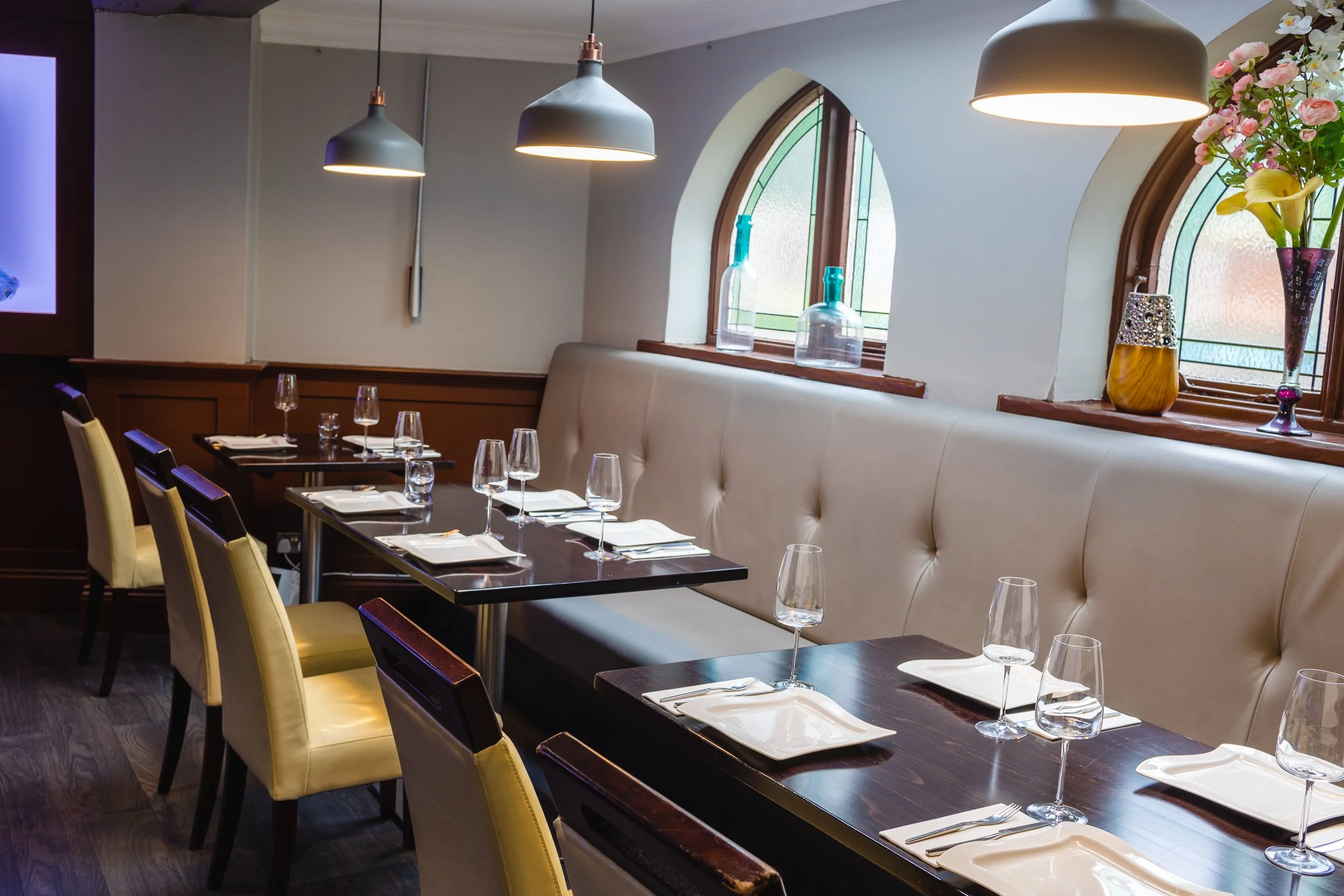 Upscale restaurant with dark wooden tables set with white napkins, wine glasses, and white plates, beige upholstered banquette, and yellow and purple chairs. French windows with stained glass and floral vases with flowers and bottles.