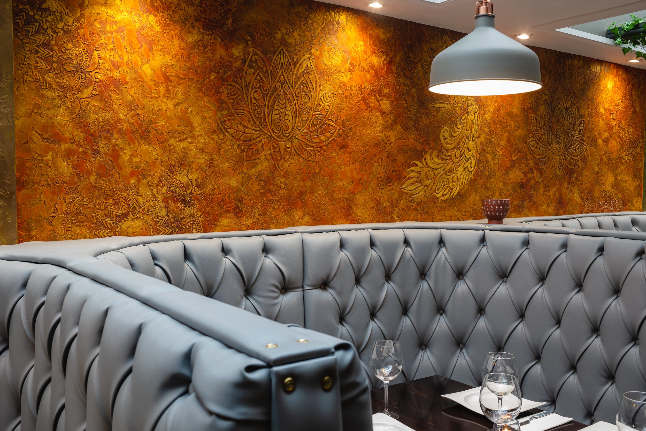 Interior of a restaurant with tufted gray seating, an orange and gold patterned wall with intricate designs, a large hanging white light, and a small vase on the table with wine glasses and tableware.