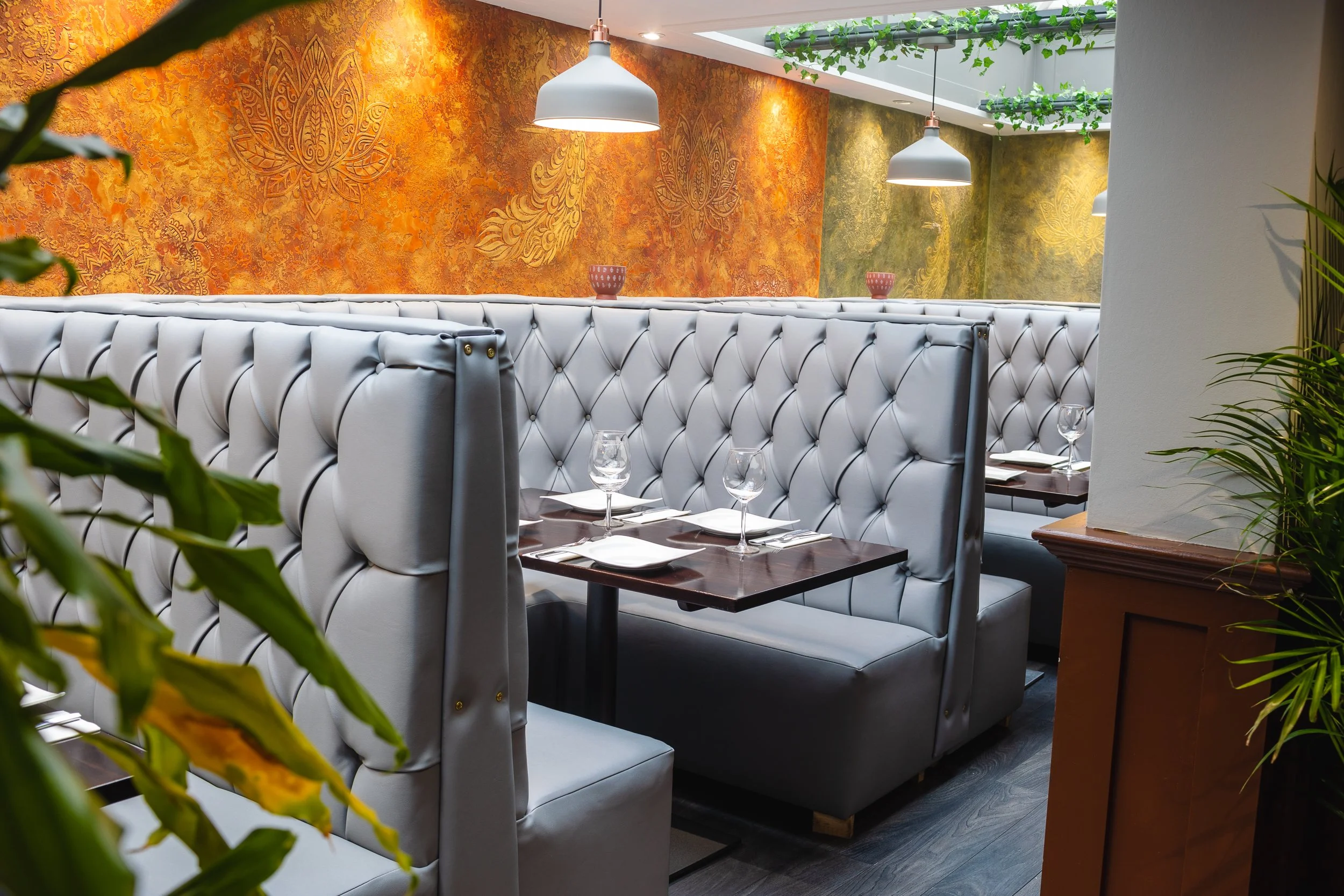 Empty restaurant booth with white tufted seating, dark wood tables set with wine glasses and napkins, colorful textured walls, hanging white pendant lights, and a skylight with green vines hanging down.