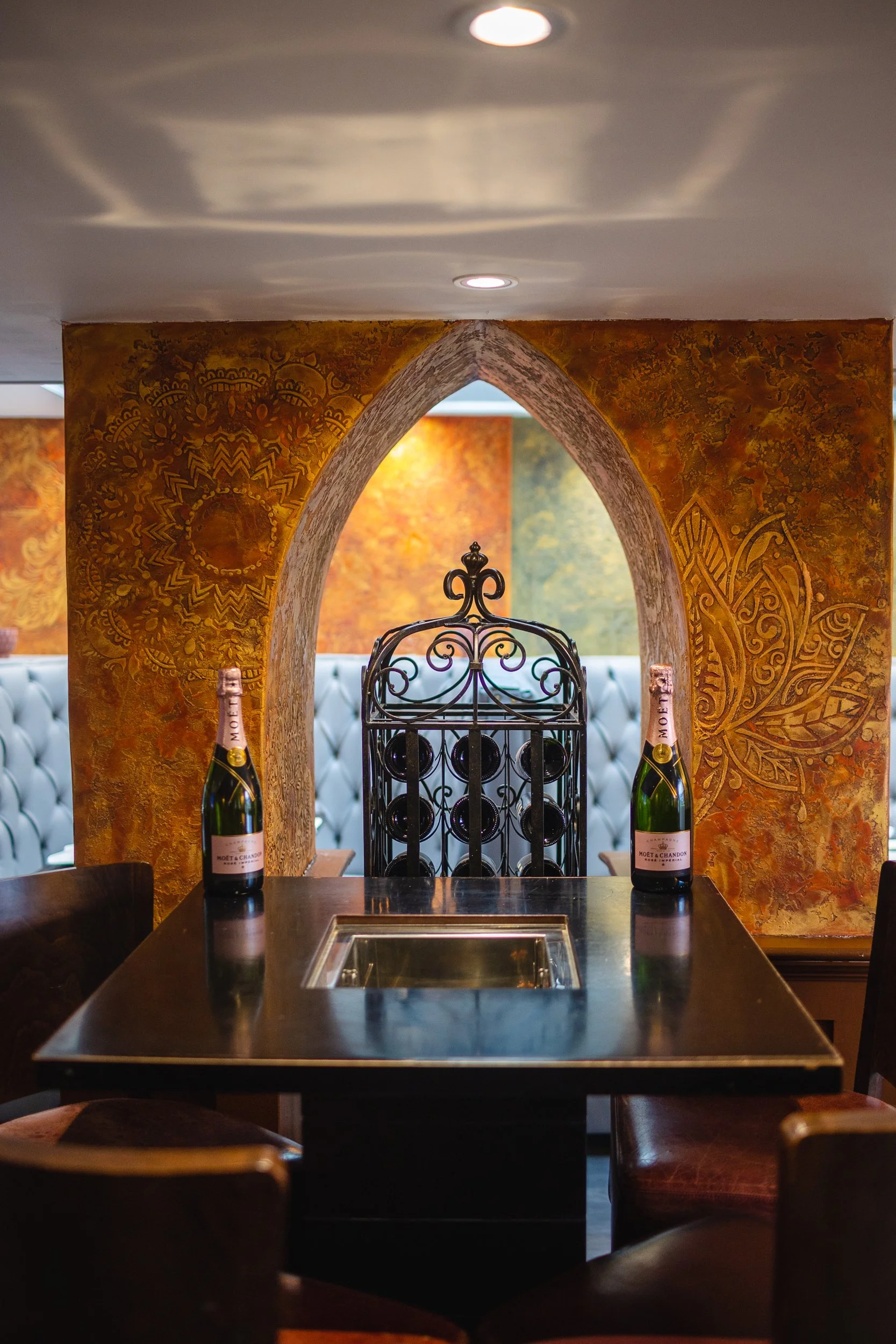 A restaurant interior featuring a black table with a built-in grill, two bottles of Moët & Chandon champagne, a decorative metal wine rack, and an ornate arched wall with warm-colored murals.