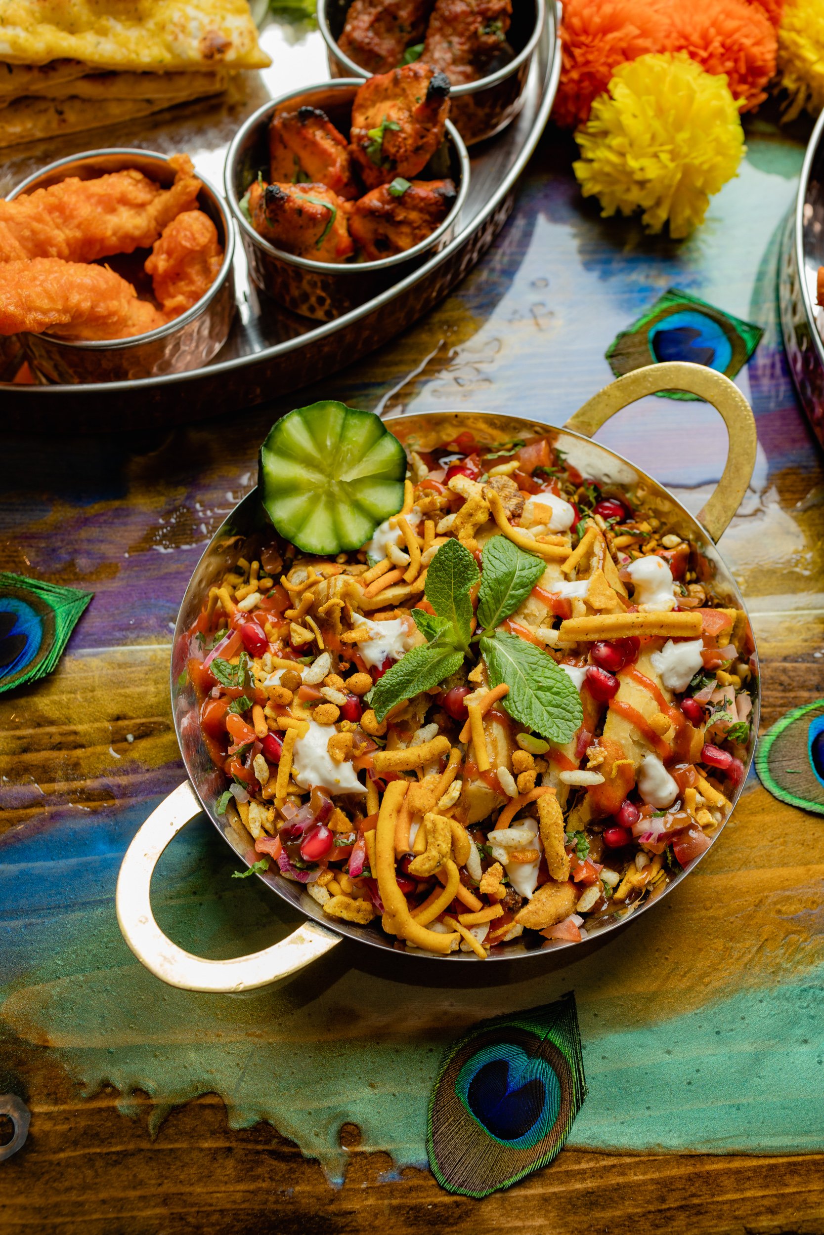 A bowl of chaat garnished with mint leaves, cucumber, pomegranate seeds, crispy sev, and yogurt on a colorful wooden table with decorative peacock feather designs.