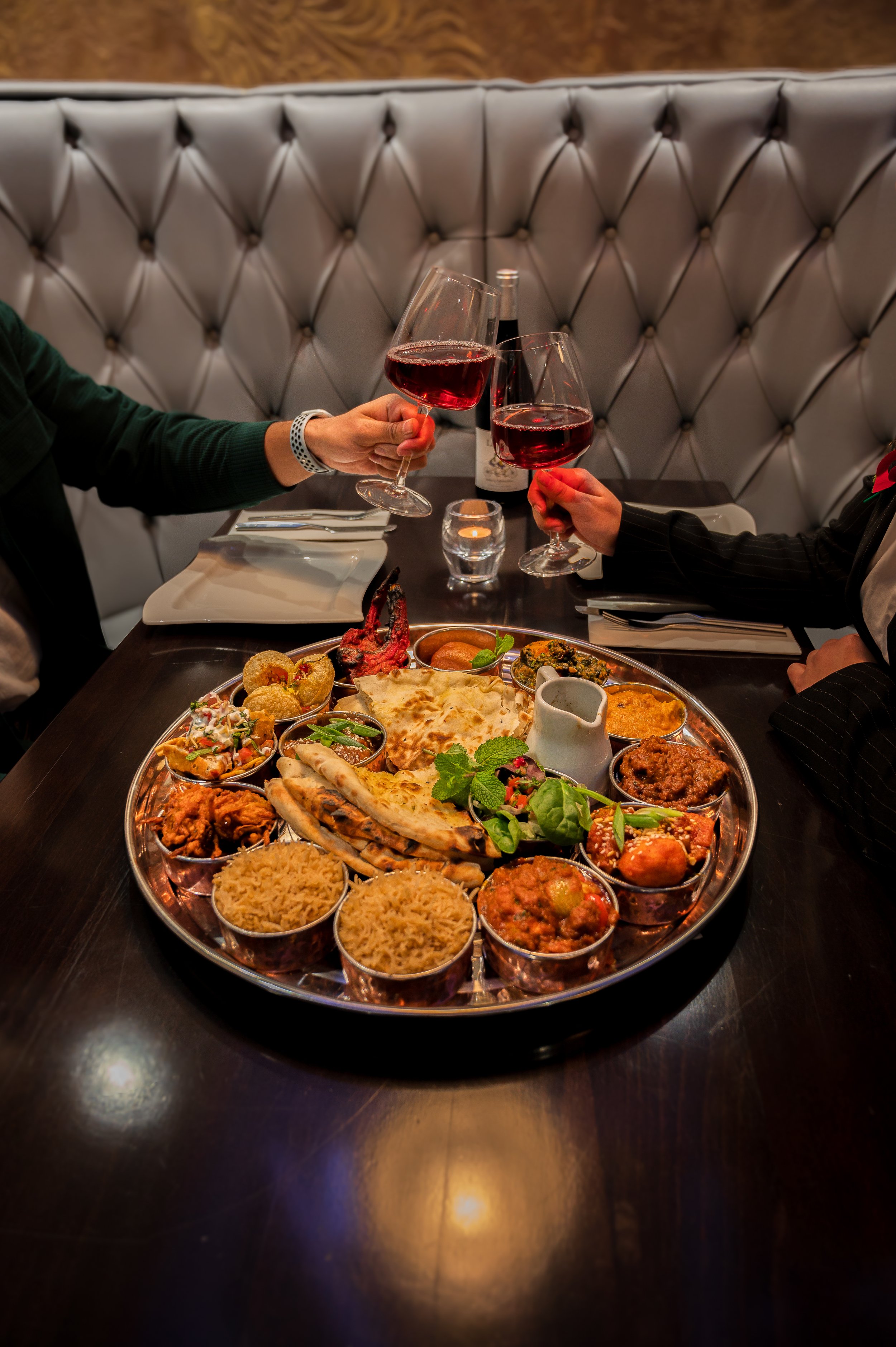 Two people clinking glasses of red wine over a large Indian thali meal at a restaurant table.