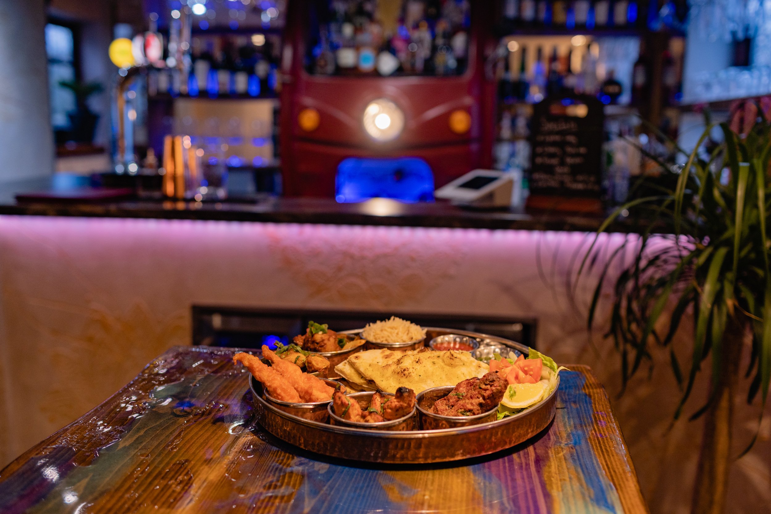 A round metal tray with Indian food including rice, naan, and various curries on a wooden table in a restaurant with a bar in the background.