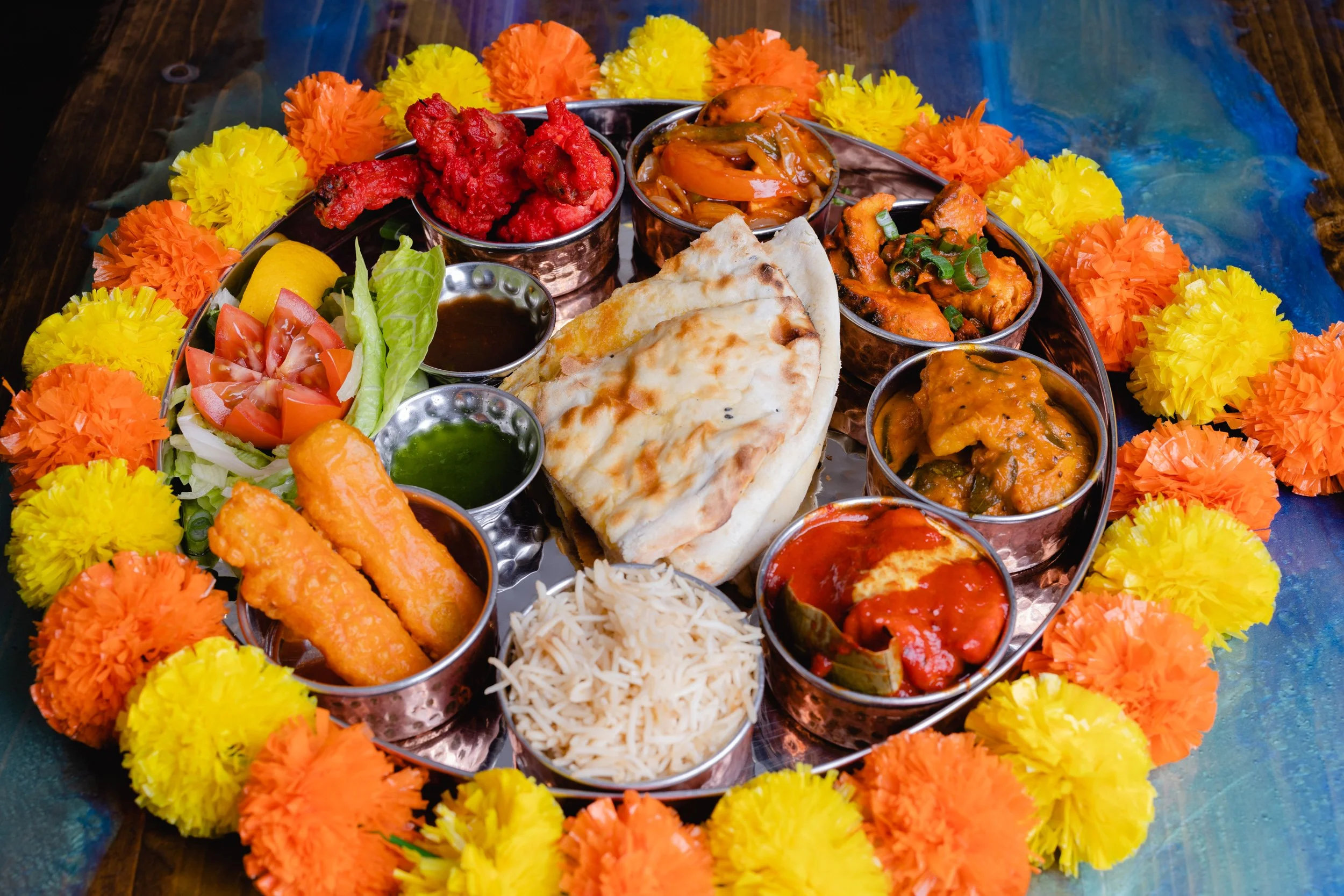 An Indian thali with various dishes, including naan bread, rice, vegetables, curries, and fried items, garnished with marigold and other flower decorations.